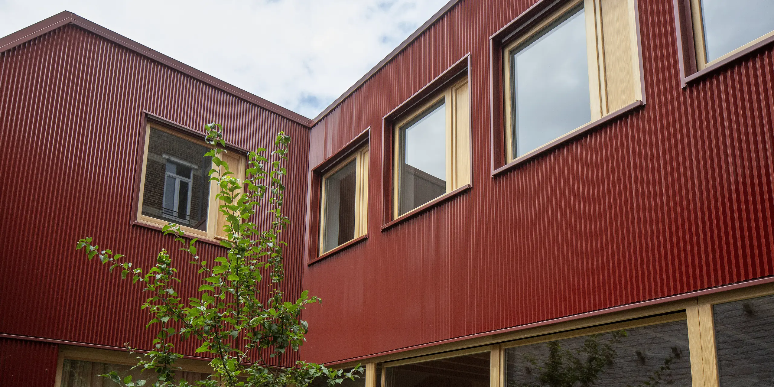 La cour intérieure rouge de Lille impressionne par le jeu dynamique des profils verticaux des poutres PREFA, du bois chaleureux et des grandes fenêtres. La couleur rouge oxyde intense crée un contraste harmonieux avec le vert frais des plantes et constitue un élément décoratif durable et sans entretien, à fort impact architectural.
