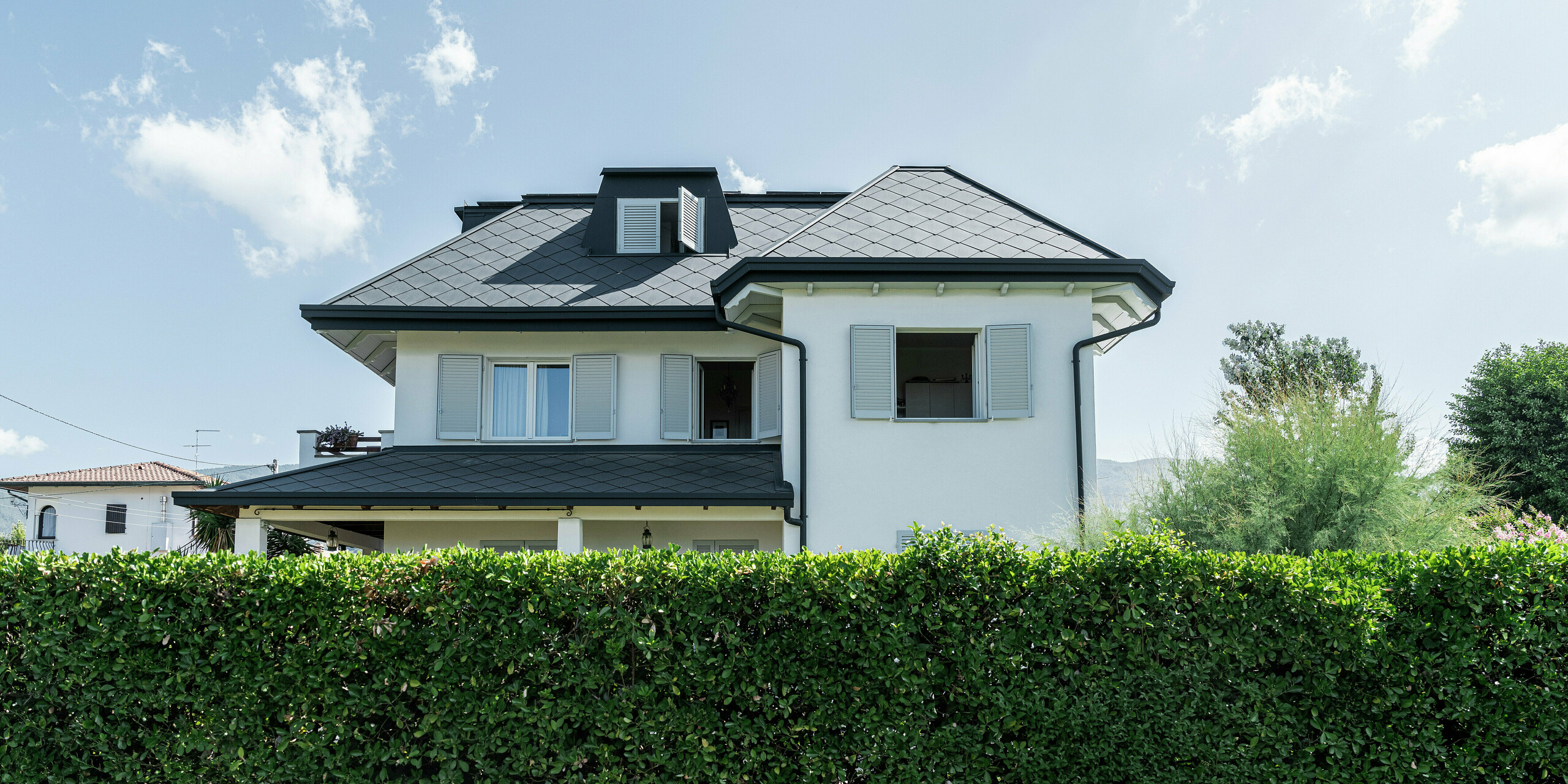 Vue plongeante sur la haie verte luxuriante d'une élégante maison individuelle à Montignoso, en Italie. Dans la partie supérieure de l'image, la couverture du toit avec le losange de toiture 44 × 44  PREFA de haute qualité en P.10 gris pierre attire particulièrement l'attention. Le toit en aluminium confère au bâtiment une apparence moderne et élégante et constitue non seulement un point fort du design, mais également une résistance extrême aux tempêtes, à la corrosion et aux intempéries. La combinaison d'une façade claire, d'un toit sombre et d'un environnement verdoyant crée une image globale harmonieuse.