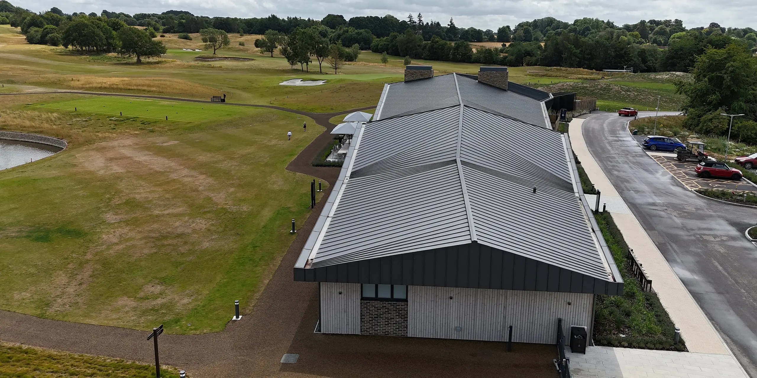 L'élégante toiture à joints debout PREFALZ du Basingstoke Golf Club, dans le Hampshire, avec ses courbes harmonieuses, attire tous les regards. La toiture gris sombre P.10 apporte des touches de couleur saisissantes qui se fondent parfaitement dans le vaste paysage du parcours de golf. Au premier plan, le pignon du bâtiment et le terrain de golf impeccablement entretenu se dessinent, complétant à merveille l'architecture moderne du club-house.