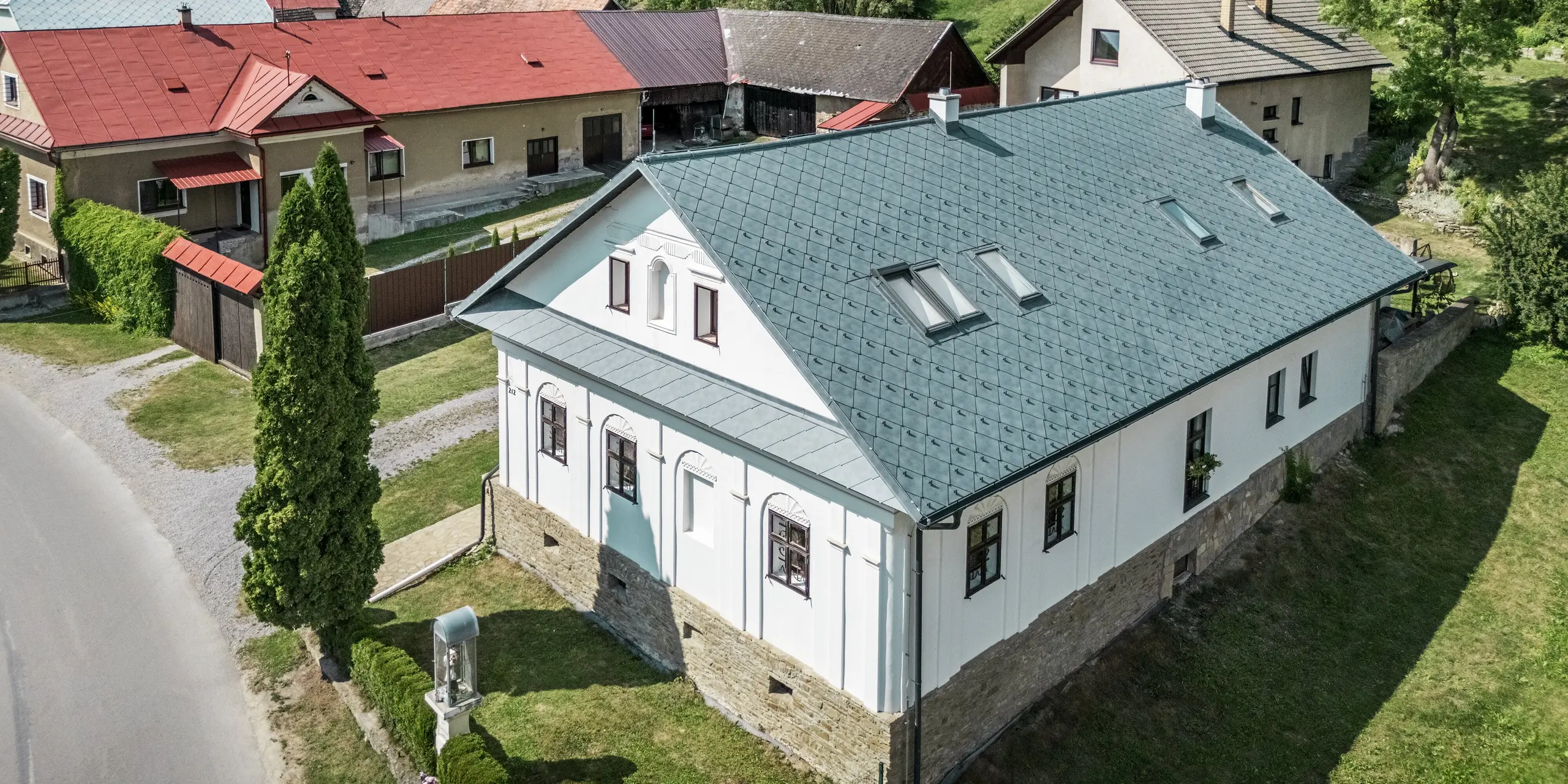 L'image présente une maison d'artisan rénovée à Klin, en Slovaquie, avec un toit à pignon recouvert de losanges PREFA 44 x 44 couleur Gris Pierre P.10. La charpente symétrique, intégrant des puits de lumière, apporte non seulement une touche esthétique, mais aussi une abondante lumière naturelle à l'intérieur de la maison. La toiture en aluminium s'harmonise parfaitement avec la façade classique en pierre naturelle et offre une excellente durabilité, une résistance à la corrosion et une grande légèreté. Cette solution de toiture durable est idéale pour la rénovation de bâtiments historiques, car elle garantit une fonctionnalité optimale et une protection efficace contre les intempéries.