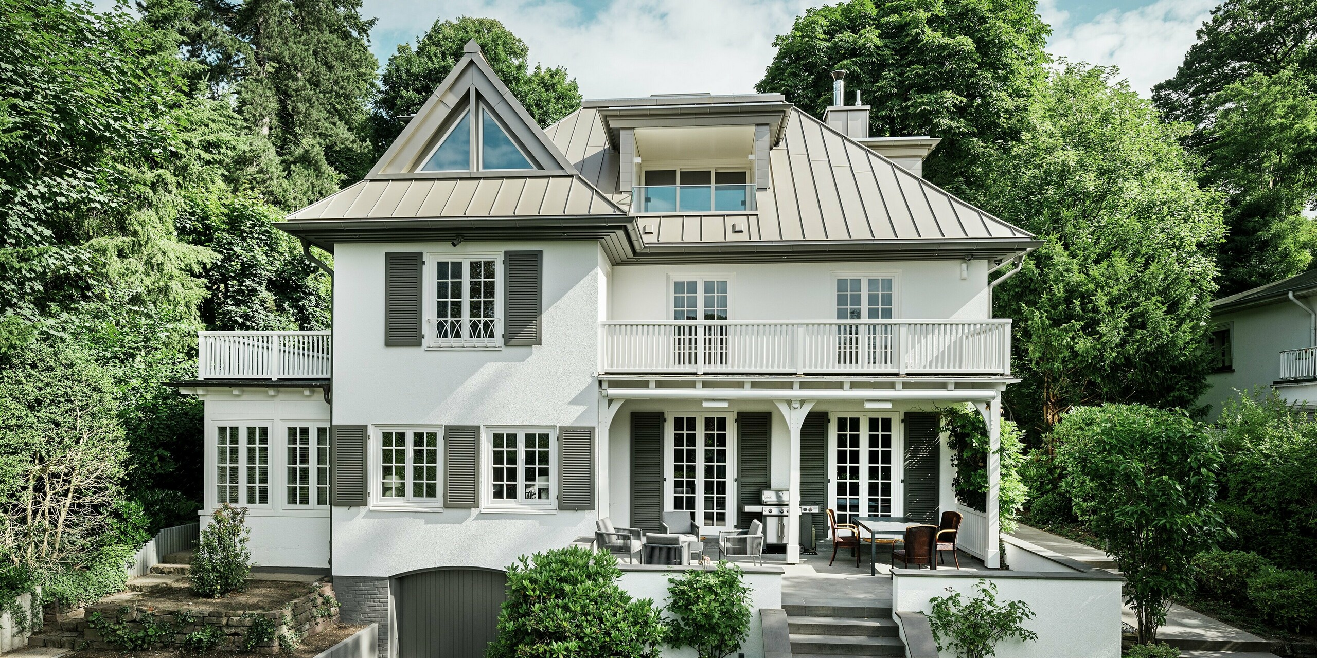 L'image présente la Villa Blankenese à Hambourg, vue de face, nichée dans un écrin de verdure. La remarquable toiture PREFALZ en bronze souligne l'architecture et confère au bâtiment une élégance intemporelle. La technique précise des joints debout garantit des lignes épurées et une résistance optimale aux intempéries. Des bardages PREFA couleur bronze habillent les lucarnes et les éléments de façade, accentuant ainsi l'harmonie entre la toiture et la façade.