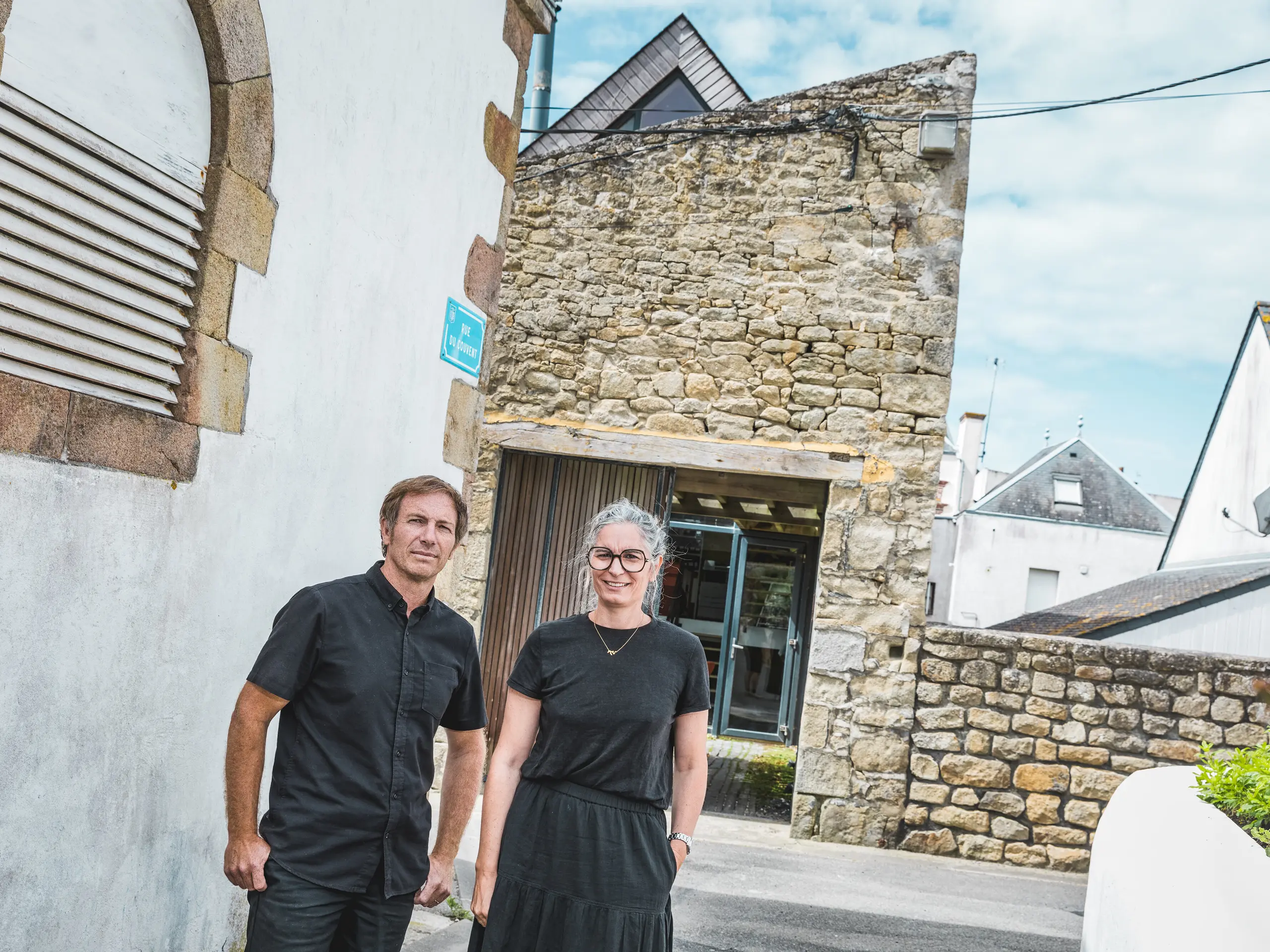 Dans une ruelle bretonne étroite, les deux architectes Anne-Charlotte et Jean-François Riguidel se tiennent devant une façade en pierre naturelle remarquable. Tous deux sont vêtus de noir. Le contraste saisissant entre la maçonnerie traditionnelle, les façades blanches et l'architecture moderne de la toiture met en lumière la fusion réussie des traditions architecturales régionales et du design contemporain.