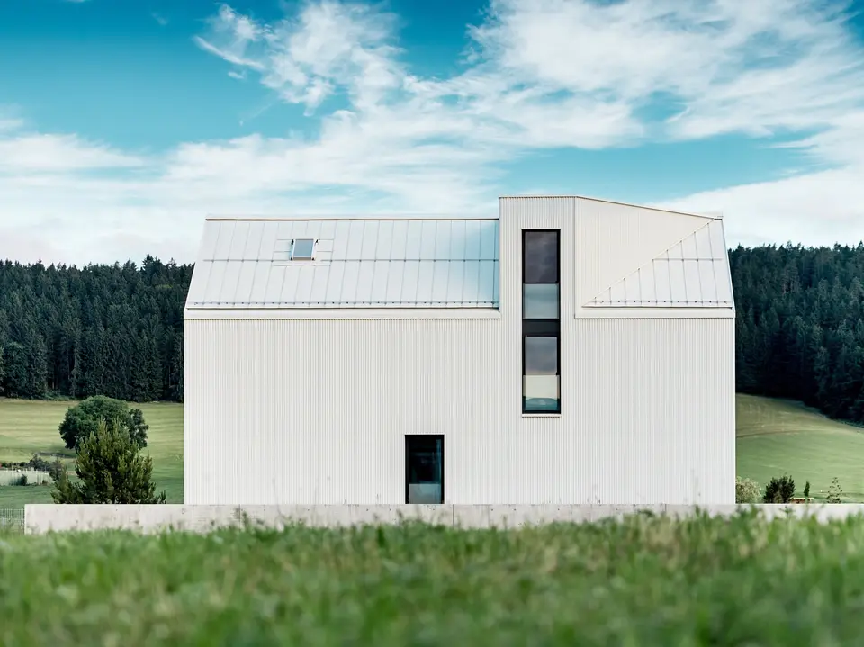 L'image présente une maison individuelle à Freistadt, vue de profil, mettant en valeur son architecture minimaliste et remarquable. La façade en aluminium blanc pur PREFALZ et le toit à pignon abrupt dessinent une silhouette épurée et moderne qui s'intègre harmonieusement à la verdure environnante. Les grandes fenêtres et l'élégante lucarne soulignent la fonctionnalité du design, tandis que la qualité de l'aluminium garantit à la fois une esthétique durable et une excellente résistance aux intempéries. Par sa forme simple et l'utilisation de matériaux pérennes, le bâtiment dégage une élégance intemporelle et représente une parfaite harmonie entre architecture et nature.