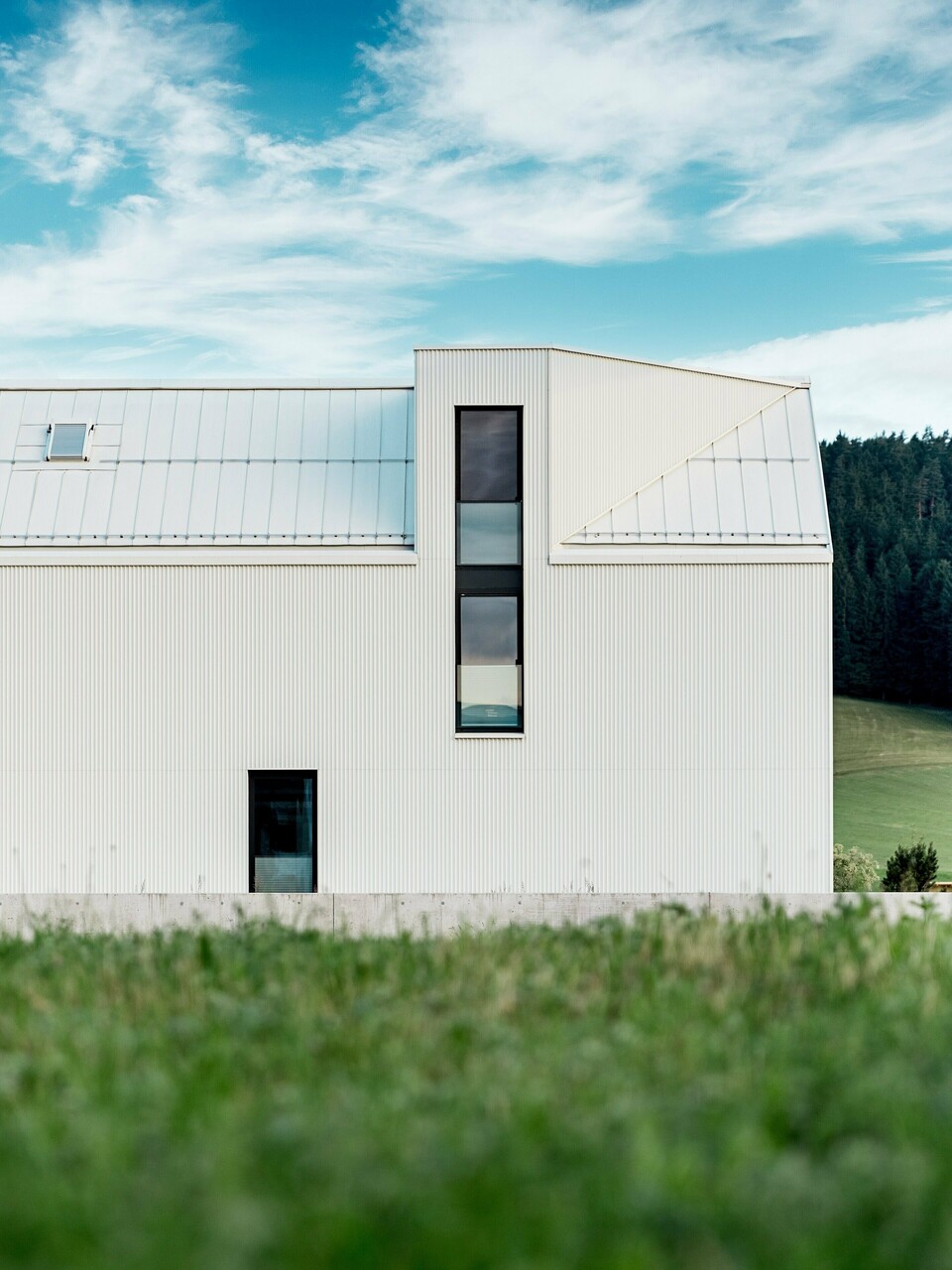 L'image présente une maison individuelle à Freistadt, vue de profil, mettant en valeur son architecture minimaliste et remarquable. La façade en aluminium blanc pur PREFALZ et le toit à pignon abrupt dessinent une silhouette épurée et moderne qui s'intègre harmonieusement à la verdure environnante. Les grandes fenêtres et l'élégante lucarne soulignent la fonctionnalité du design, tandis que la qualité de l'aluminium garantit à la fois une esthétique durable et une excellente résistance aux intempéries. Par sa forme simple et l'utilisation de matériaux pérennes, le bâtiment dégage une élégance intemporelle et représente une parfaite harmonie entre architecture et nature.