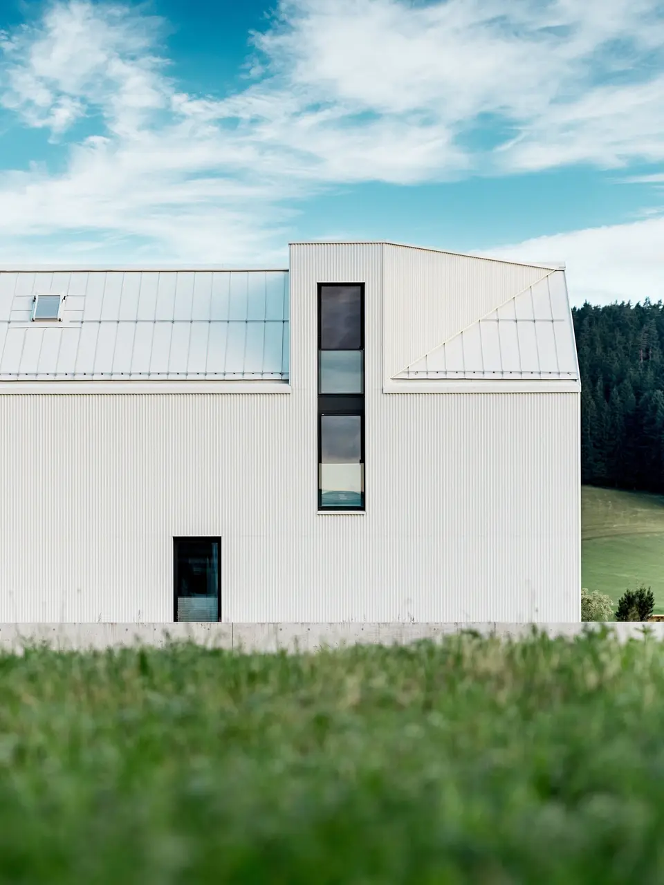 L'image présente une maison individuelle à Freistadt, vue de profil, mettant en valeur son architecture minimaliste et remarquable. La façade en aluminium blanc pur PREFALZ et le toit à pignon abrupt dessinent une silhouette épurée et moderne qui s'intègre harmonieusement à la verdure environnante. Les grandes fenêtres et l'élégante lucarne soulignent la fonctionnalité du design, tandis que la qualité de l'aluminium garantit à la fois une esthétique durable et une excellente résistance aux intempéries. Par sa forme simple et l'utilisation de matériaux pérennes, le bâtiment dégage une élégance intemporelle et représente une parfaite harmonie entre architecture et nature.