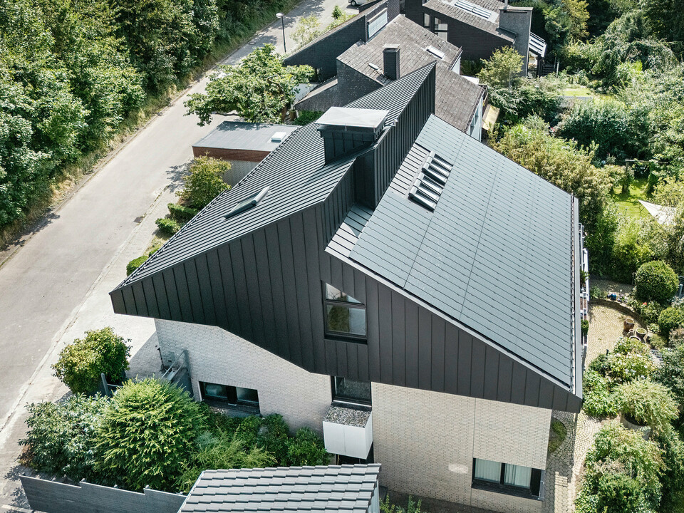 Vue oblique d'une maison unifamiliale moderne à Meschede, en Allemagne. Le toit en pente décalé a été revêtu de PREFALZ en P.10 noir. Le toit en aluminium constitue un élément visuel marquant et offre au bâtiment une protection permanente. La toiture à double joint debout de précision s'étend du toit à travers la façade jusqu'aux pignons. Entouré d'une végétation luxuriante et d'une rue calme, le bâtiment économe en énergie avec les modules solaires innovants PREFALZ s'intègre parfaitement dans l'environnement de vie naturel.