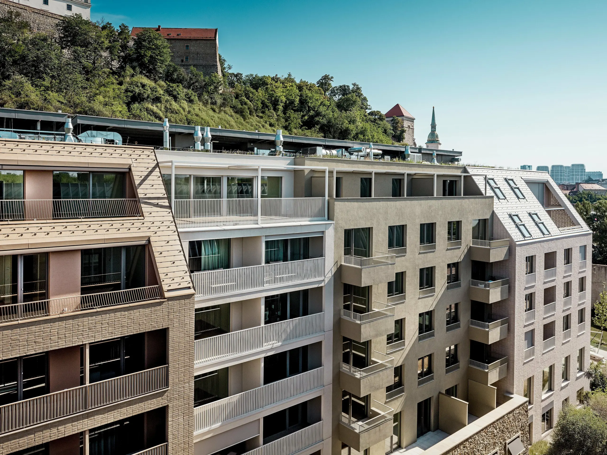 Un complexe résidentiel urbain du quartier Vydrica à Bratislava, offrant un contraste saisissant entre architecture moderne et cadre historique à proximité du château : les toitures pentues, aux lignes épurées, ornées de lucarnes et de vastes terrasses en aluminium PREFA brun sable et blanc, constituent les points forts visuels. Ces solutions en aluminium de haute qualité permettent des formes de toiture complexes, impressionnent par leur durabilité, leur résistance aux intempéries et leur faible besoin d’entretien, et créent une enveloppe de bâtiment durable et esthétiquement sophistiquée en milieu urbain.