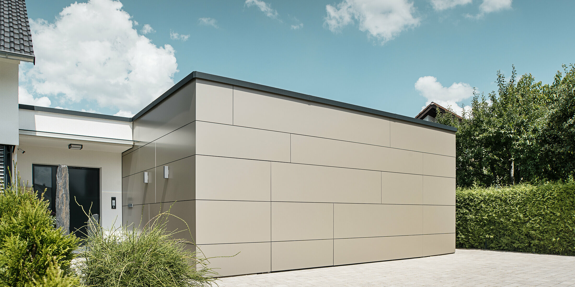 Gesamtansicht einer Garage im geschlossenen Zustand. Das geschlossene Garagentor lässt die Öffnungsmöglichkeit nur schwer erkennen, da die PREFABOND Aluminium Verbundplatten in Bronze das Gebäude fast nahtlos verkleiden. Die Pflanzen vor der Eingangstür zeugen von der Pflege des schönen Einfamilienhauses.