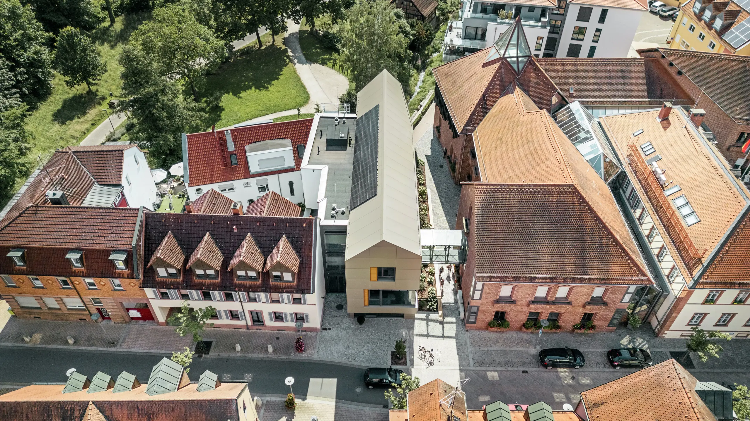 La mairie d'Alzenau, en Allemagne, est située au centre d'un paysage urbain historique, entourée de bâtiments traditionnels aux toits de tuiles rouges. La mairie moderne contraste avec ces bâtiments, sa façade étant revêtue de panneaux composites en aluminium PREFA dans un élégant ton bronze. L'architecture innovante se démarque clairement des bâtiments classiques voisins tout en s'intégrant harmonieusement dans le paysage urbain.