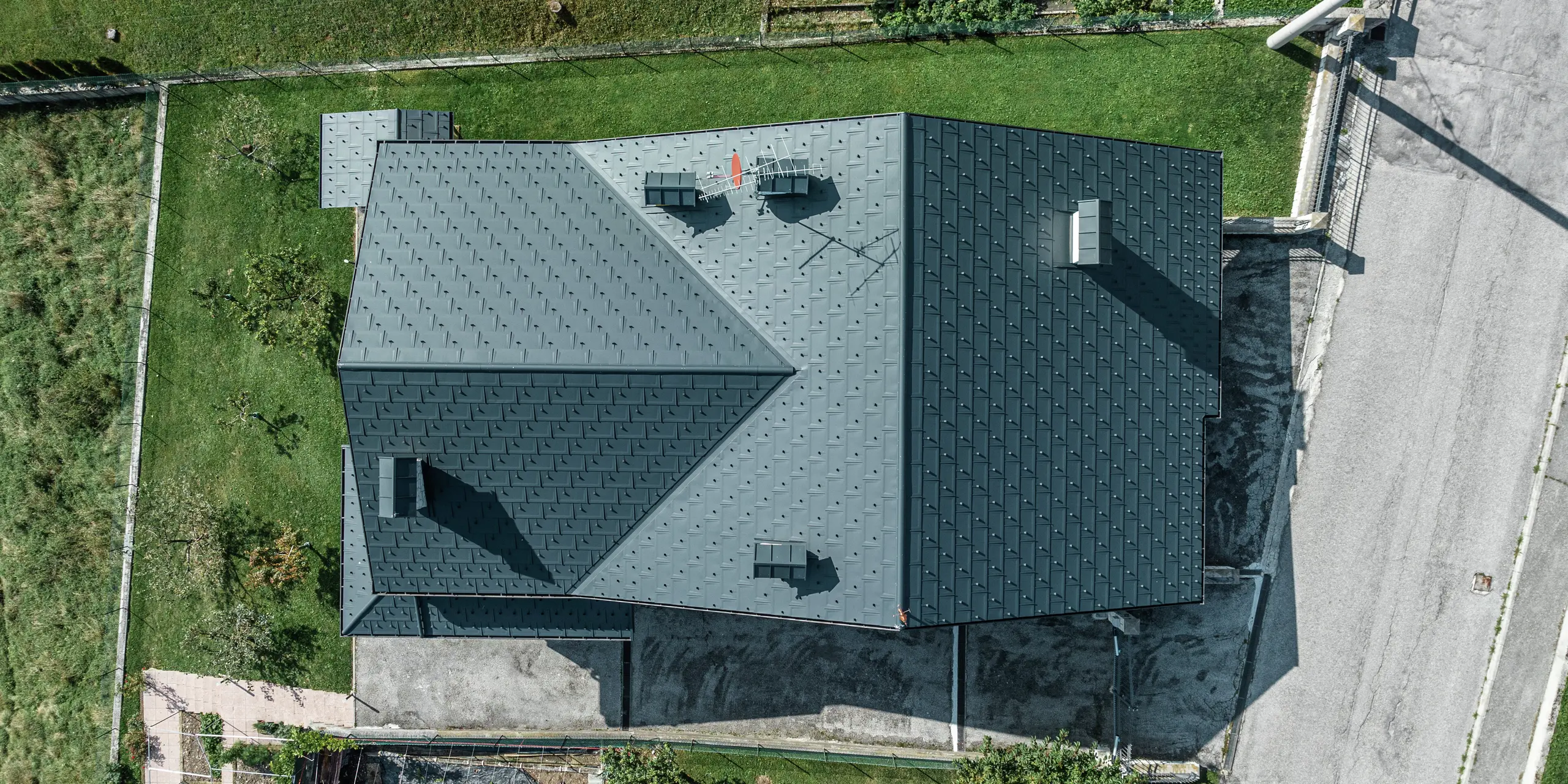 Prise de vue par drone d'une maison familiale à Gallio avec une couverture de toit PREFA de haute qualité composée de panneaux de toiture R.16 en P.10 gris sombre. La solution de toiture en aluminium impressionne par son aspect moderne, sa résistance extrême aux intempéries et sa durabilité. Les crochets pare-neige offrent une sécurité supplémentaire dans les régions alpines. La finition précise et le système de drainage sophistiqué garantissent une protection optimale contre la pluie et la neige. Un toit nécessitant peu d’entretien pour des générations.