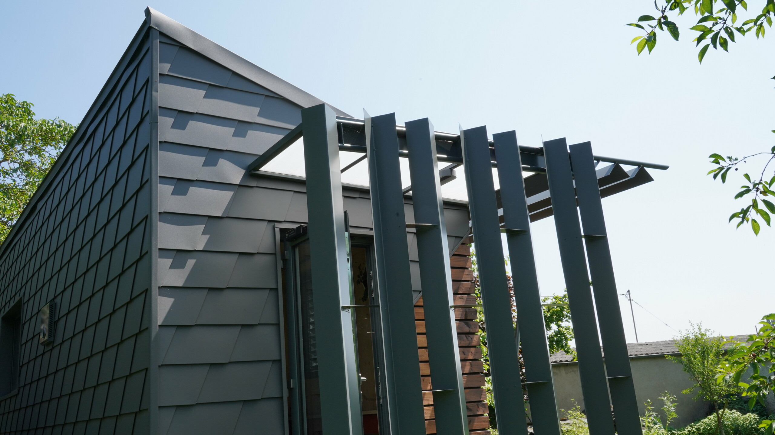 Zoom sur l'entrée de ce bureau mobile. La façade est partiellement recouverte de bois et de bardeau en aluminium PREFA. L'entrée est couverte des intempéries grâce à un jeu de menuiserie en aluminium.