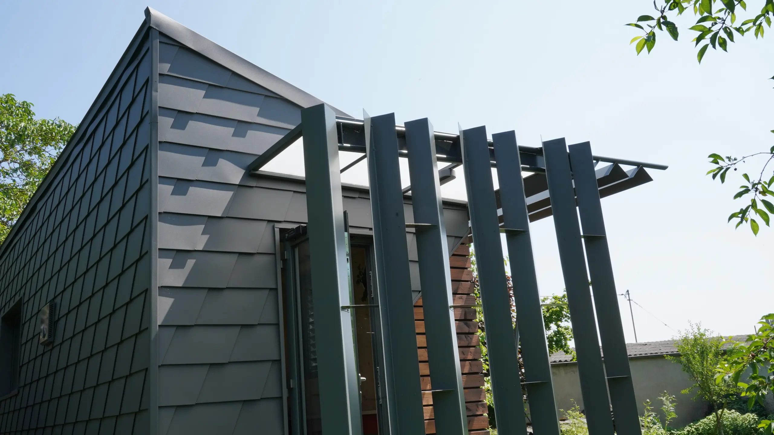 Zoom sur l'entrée de ce bureau mobile. La façade est partiellement recouverte de bois et de bardeau en aluminium PREFA. L'entrée est couverte des intempéries grâce à un jeu de menuiserie en aluminium.