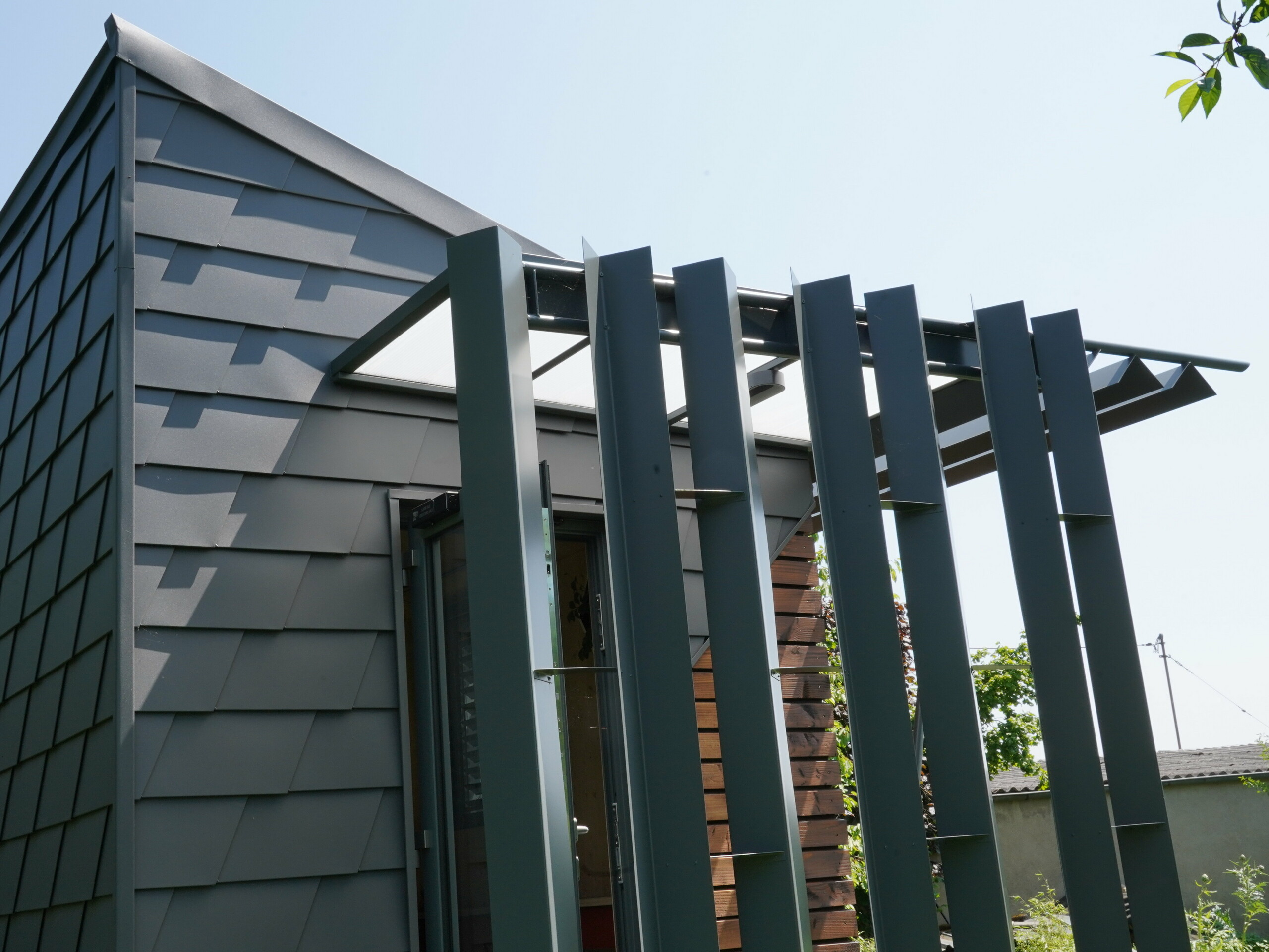 Zoom sur l'entrée de ce bureau mobile. La façade est partiellement recouverte de bois et de bardeau en aluminium PREFA. L'entrée est couverte des intempéries grâce à un jeu de menuiserie en aluminium.