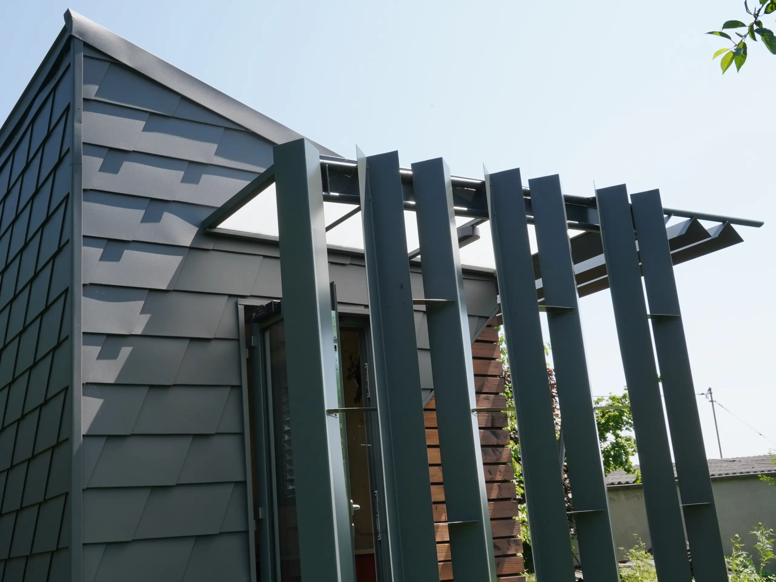 Zoom sur l'entrée de ce bureau mobile. La façade est partiellement recouverte de bois et de bardeau en aluminium PREFA. L'entrée est couverte des intempéries grâce à un jeu de menuiserie en aluminium.