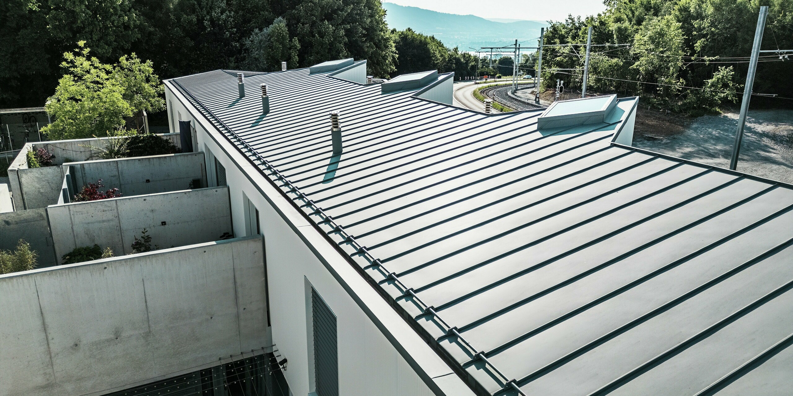 Vue en perspective d'un toit allongé d'un ensemble de mini-maisons à Zollikerberg, doté d'une toiture à joints debout PREFALZ de haute qualité, couleur gris quartz. Résistante aux intempéries, la toiture en aluminium PREFA s'intègre harmonieusement à l'architecture moderne et au cadre naturel. Ses lignes épurées, sa charpente intégrée et le choix de matériaux durables en font une solution haut de gamme et pérenne.