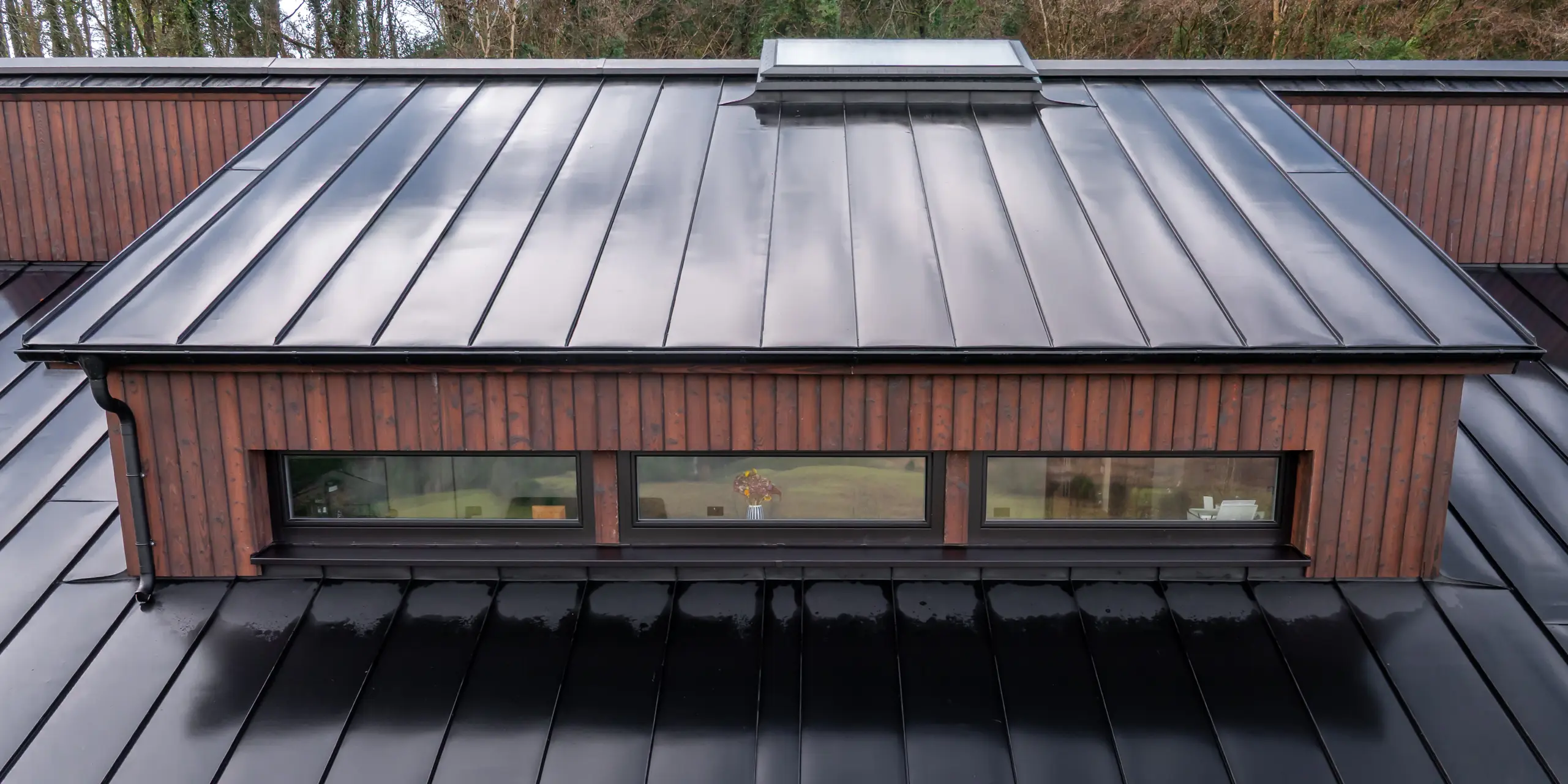 Vue de dessus de la lucarne de la Maison Holywell à Sligo : les lignes nettes de la toiture en aluminium PREFALZ P.10 Noir guident le regard le long des joints debout précis et soulignent le design minimaliste. La surface lisse et légèrement réfléchissante met en valeur la qualité de la réalisation et crée un contraste saisissant avec la façade en bois chaleureuse. Le système de toiture PREFA impressionne par sa durabilité, sa résistance à la corrosion et sa protection fiable face aux intempéries.
