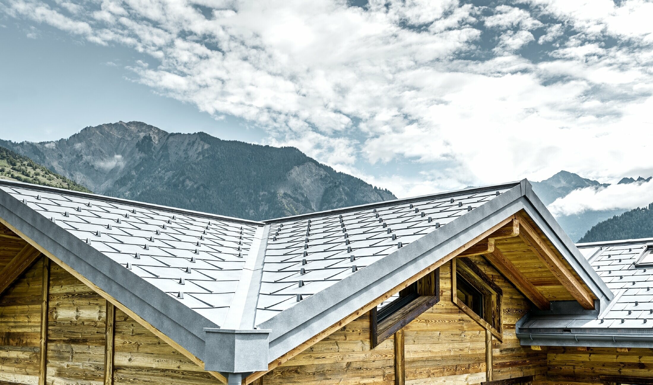 Toiture d’un chalet rustique en Suisse avec façade en bois et toiture en alu de PREFA. Le choix s’est porté sur la tuile R.16 couleur gris pierre. On peut voir la toiture anguleuse et la noue.