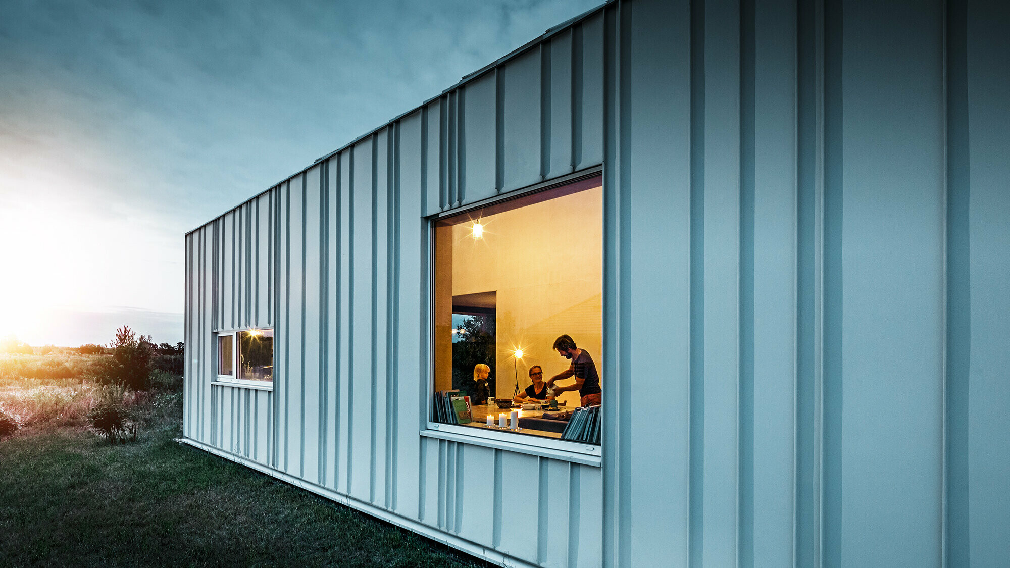 Façade avec joint debout et différentes largeurs de lames en PREFALZ de P.10 blanc PREFA photographiée au crépuscule. À travers la fenêtre de la salle à manger, on peut voir une famille en train de dîner.