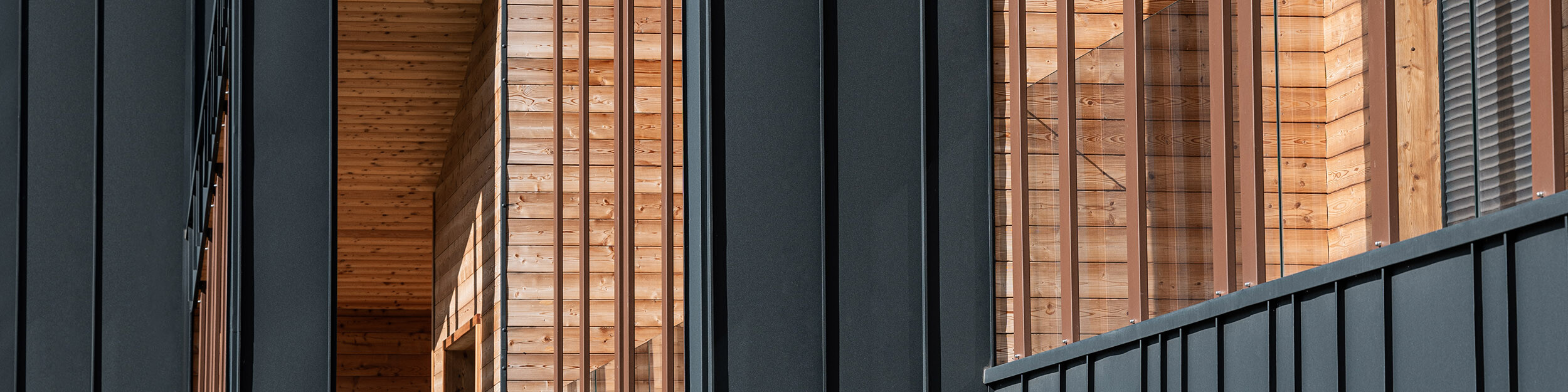 Gros plan de la façade du bâtiment moderne montrant une symbiose élégante entre des bandes de Prefalz gris foncé et des poutres en bois aux couleurs chaudes et au veinage naturel qui structurent le tout et lui donnent de la profondeur. Les poutres sont illuminées par les rayons du soleil.