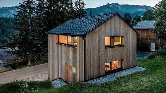 Vue depuis les hauteurs, la maison Immi se dévoile au cœur du paysage montagneux d’Ebnit. Les fenêtres chaleureusement éclairées créent un contraste saisissant avec la façade en bois naturelle et le toit solaire PREFA noir. Les panneaux solaires et les R.16 en P.10 noir, parfaitement intégrés, associent production d’énergie efficace, grande résistance et esthétique harmonieuse et contemporaine, parfaitement adaptée à l’architecture alpine.