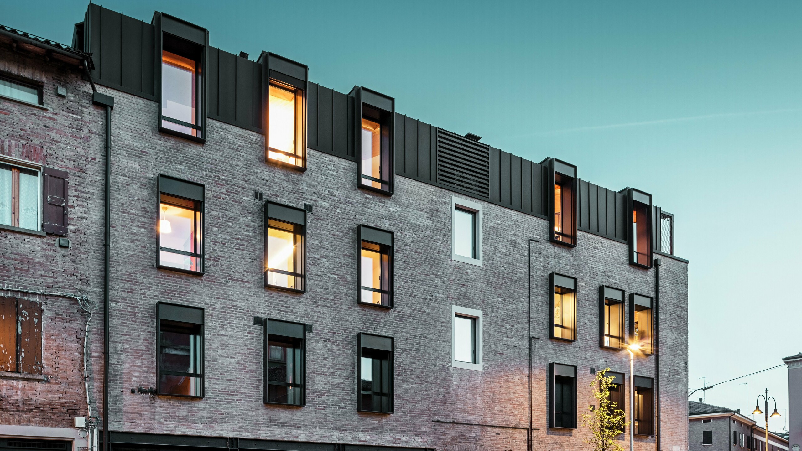 Bâtiment urbain en briques en Italie avec une toiture moderne en aluminium PREFALZ P.10 brun noisette, des fenêtres éclairées et une architecture historique environnante au crépuscule. Bâtiment urbain en briques en Italie avec toiture moderne en aluminium PREFALZ en P.10 brun noisette, fenêtres éclairées et architecture historique environnante au crépuscule.