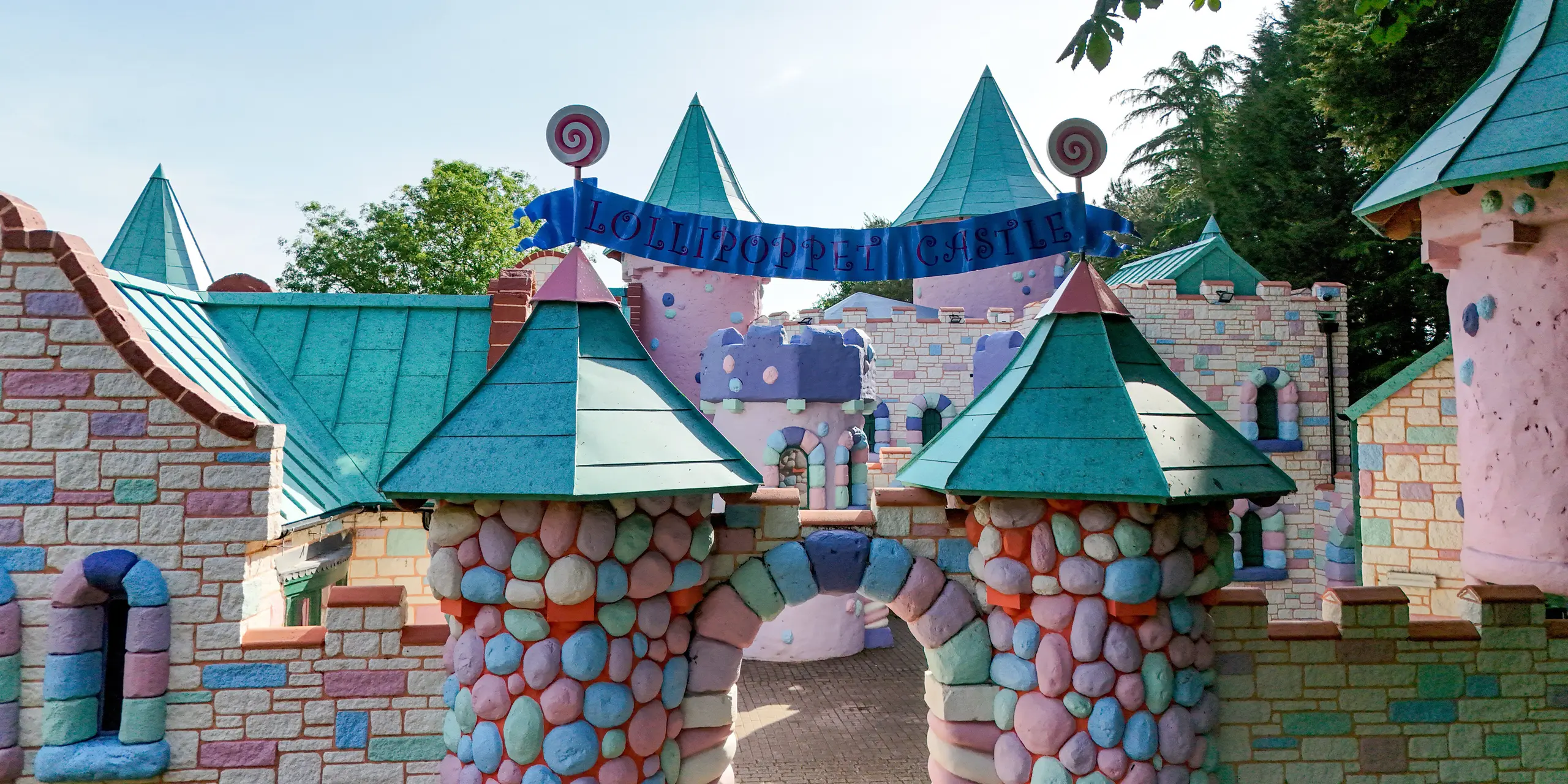 Vue de face de l'entrée du complexe féerique du Château des Sucettes à Sundown Adventureland, caractérisé par ses structures en pierre colorées, ses tours rondes ludiques et ses toits coniques remarquables qui attirent immédiatement le regard. Les toitures en aluminium PREFALZ, couleur P.10 vert de gris, définissent l'esthétique du lieu et, grâce à leurs joints debout précis, apportent une touche d'élégance et de raffinement. La complexité des formes de la toiture est élégamment mise en œuvre et souligne la liberté de conception, tandis que le système PREFA impressionne par sa durabilité, sa résistance à la corrosion et sa protection fiable par tous les temps.