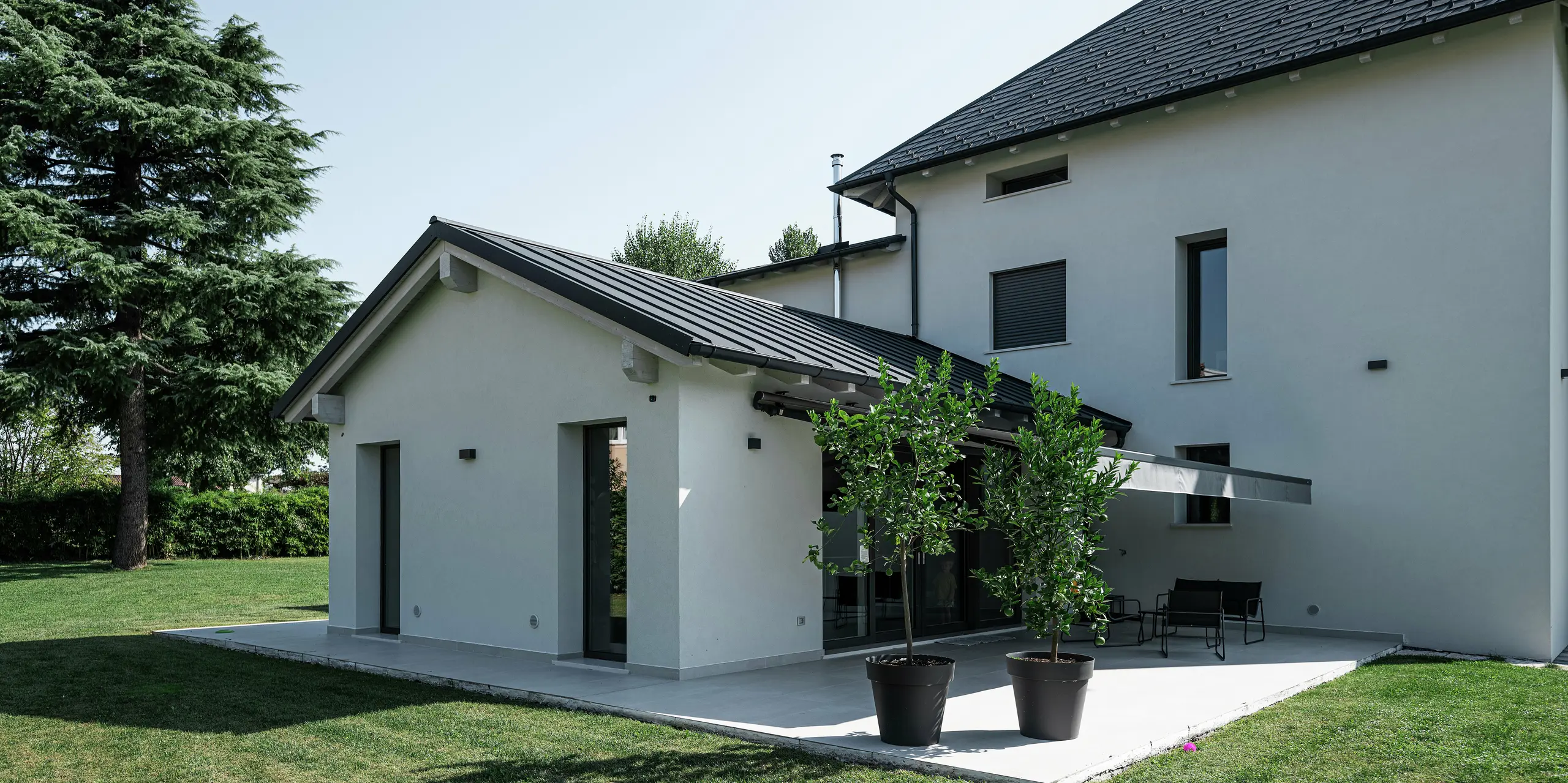 Gros plan de l'extension d'une maison familiale moderne à Marano Vicentino, en Italie, avec un revêtement de toit à joint debout PREFA en P.10 anthracite. L'élégant toit en aluminium complète harmonieusement la façade lumineuse et offre une solution durable et résistante aux intempéries. La terrasse attenante avec protection solaire crée un espace extérieur agréable dans le jardin.