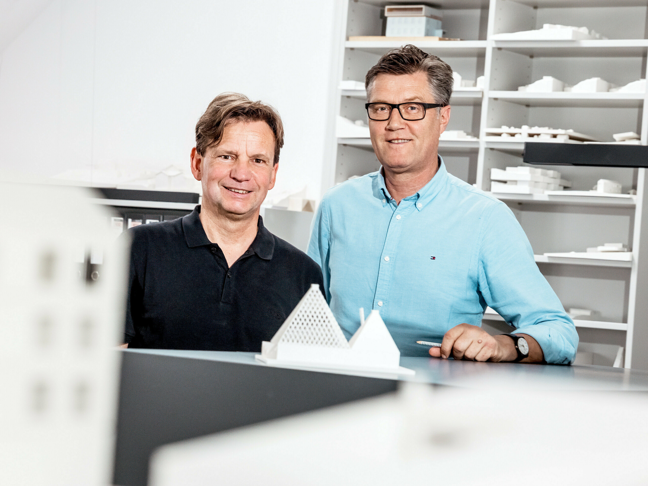  656 La photo montre les architectes de Schneider Lengauer Pühringer posant fièrement dans leur bureau devant une collection de maquettes. Ils ont conçu la maison individuelle moderne « One Shade of White » à Freistadt et sont reconnus pour leur approche novatrice et esthétique de l'architecture. Les maquettes en arrière-plan reflètent leurs concepts créatifs et témoignent de leur capacité à allier design moderne et solutions fonctionnelles. Tous deux sont vêtus de façon décontractée, soulignant l'atmosphère à la fois détendue et professionnelle de leur environnement de travail.