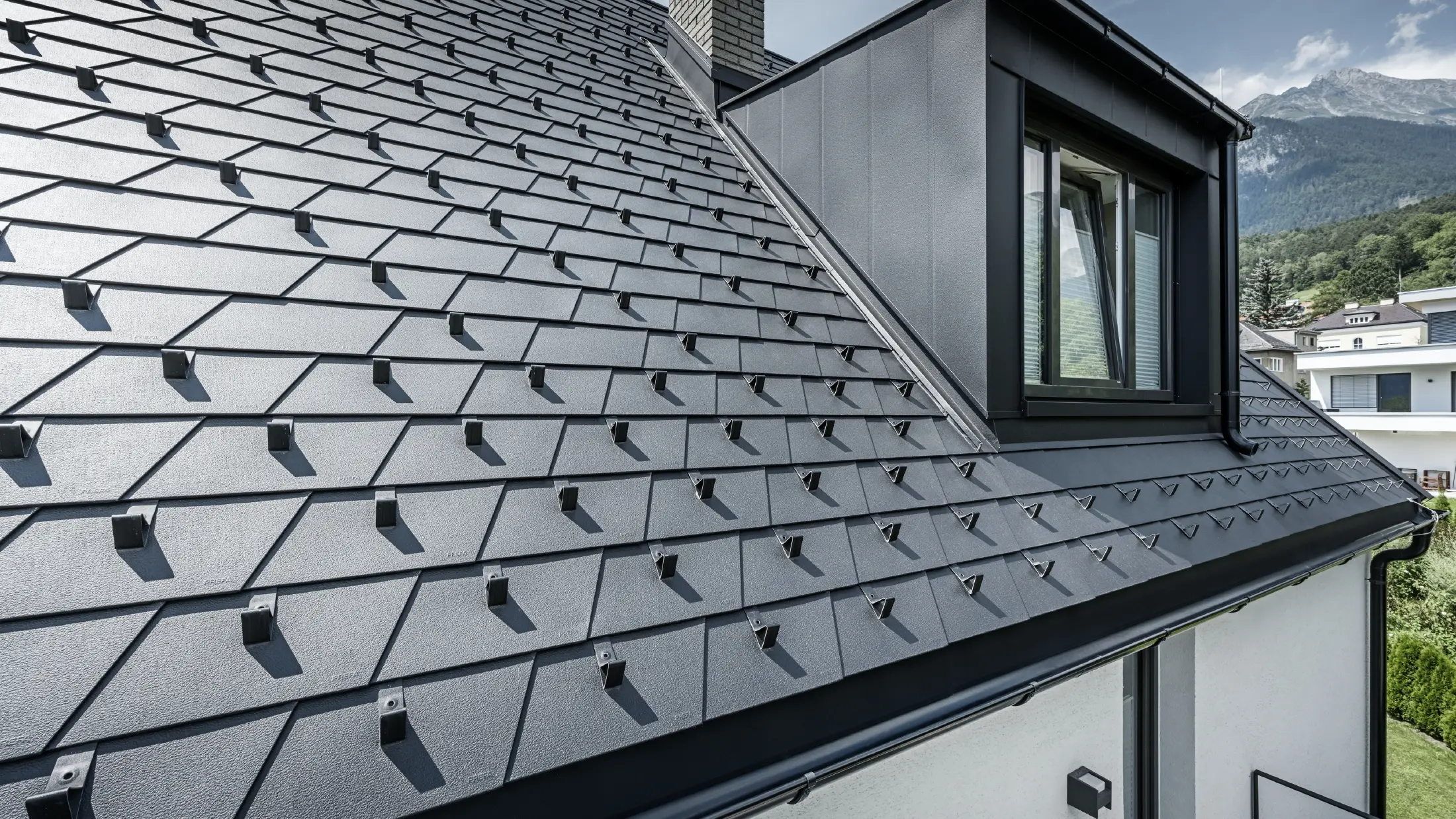 Vue détaillée d'une toiture avec lucarne et fenêtre, la surface du toit est recouverte de bardeaux PREFA noirs avec arrêts de neige.