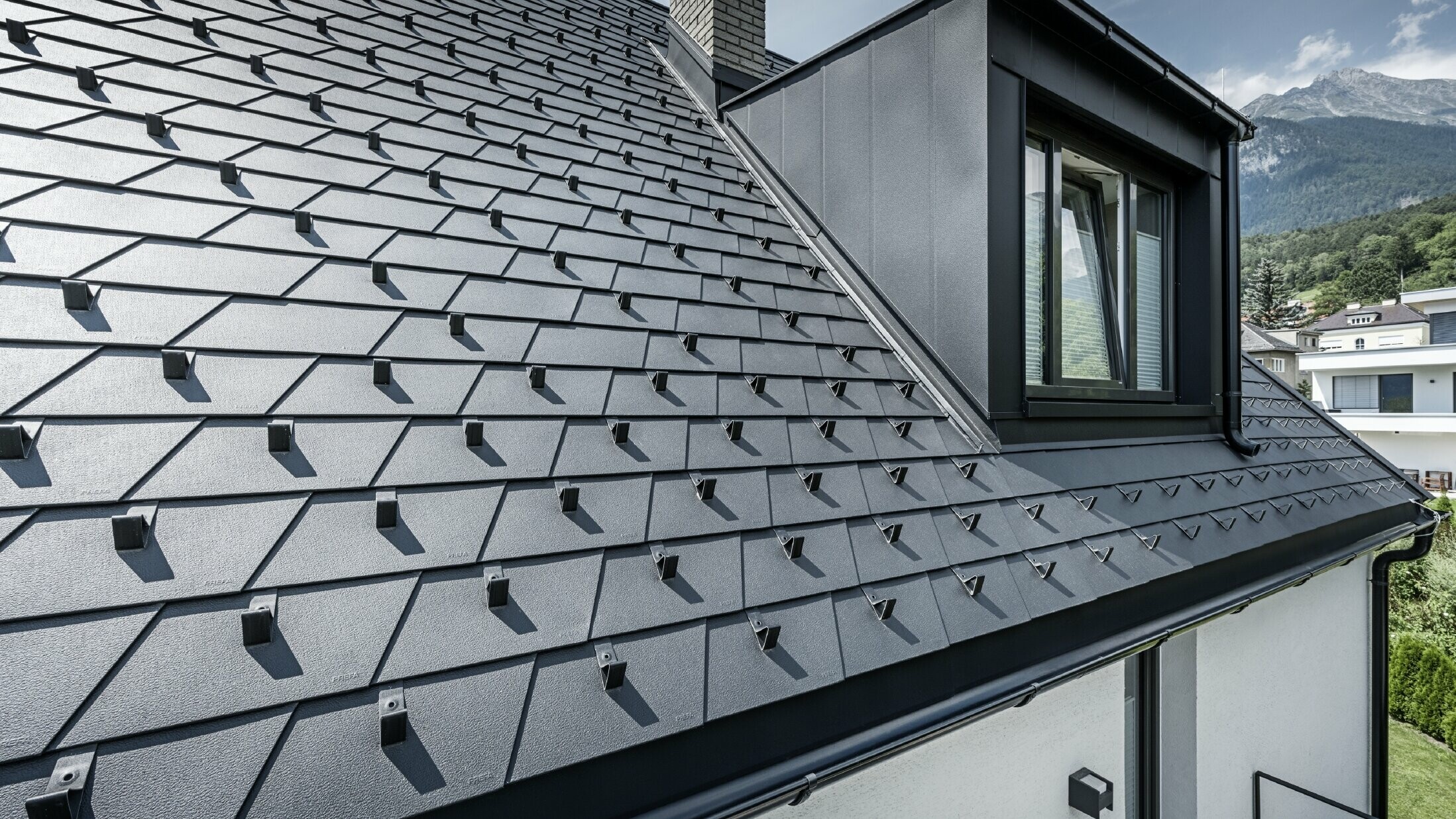 Vue détaillée d'une toiture avec lucarne et fenêtre, la surface du toit est recouverte de bardeaux PREFA noirs avec arrêts de neige.