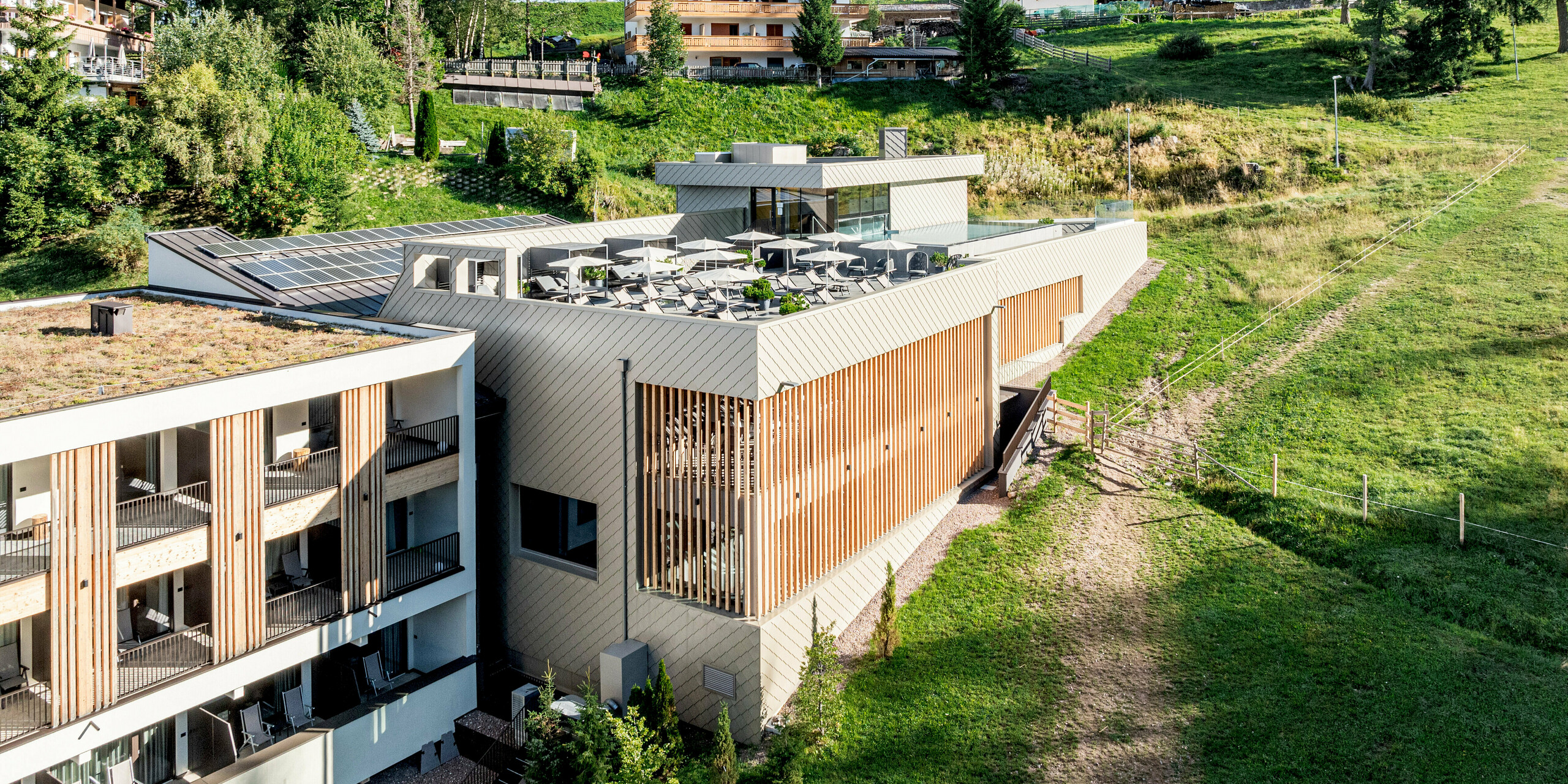 Schrägansicht des Sporthotels Obereggen in Südtirol, eingebettet in die alpine Hanglandschaft mit Wiesen und Nadelbäumen. Besonders ins Auge fällt die moderne Gebäudestruktur mit großzügiger Dachterrasse und vertikalen Holzlamellen, die mit der PREFA Aluminiumfassade harmonieren. Die Wandraute 29 × 29 in der Farbe Bronze verleiht der Fassade Tiefe und eine elegante, lebendige Anmutung. Das langlebige, witterungsbeständige und wartungsarme PREFA Aluminium eignet sich ideal für anspruchsvolle Architektur im alpinen Raum und verbindet Design mit Funktionalität.