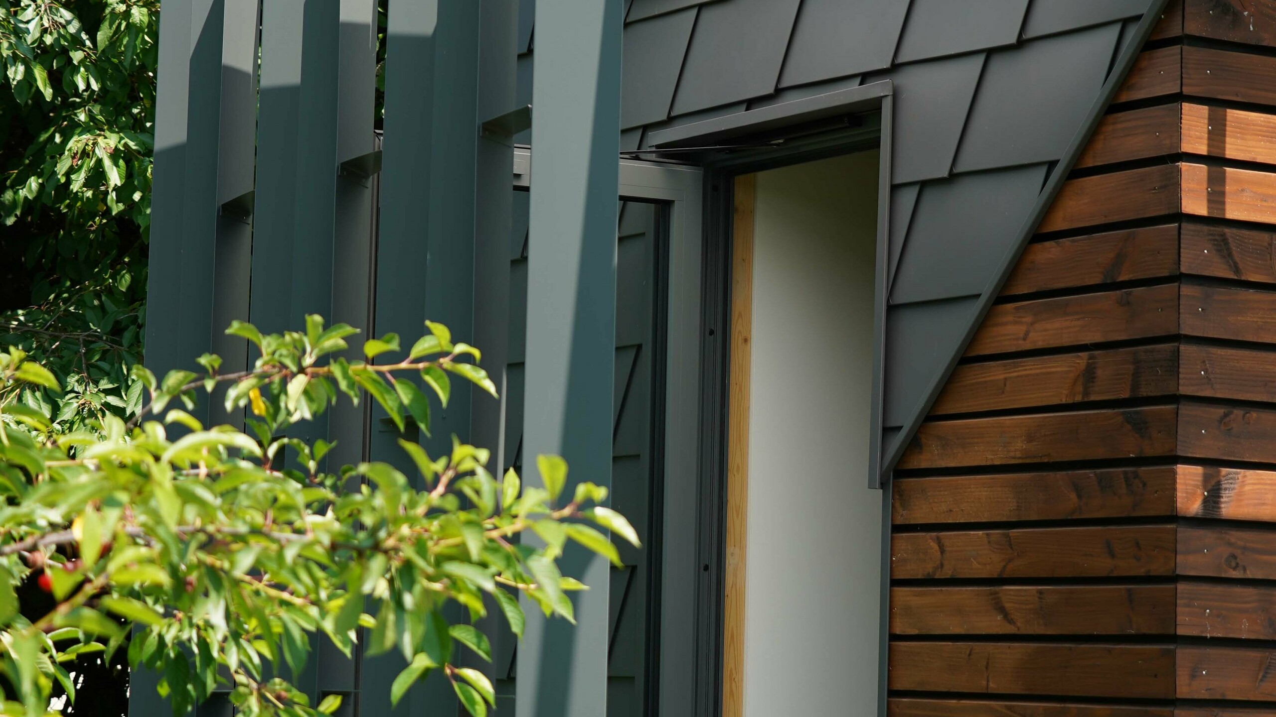 Prise de vue de la zone d'entrée de ce bureau mobile. La façade est partiellement recouverte de bois et de bardeau en aluminium PREFA. L'entrée est couverte des intempéries grâce à un jeu de menuiserie en aluminium.