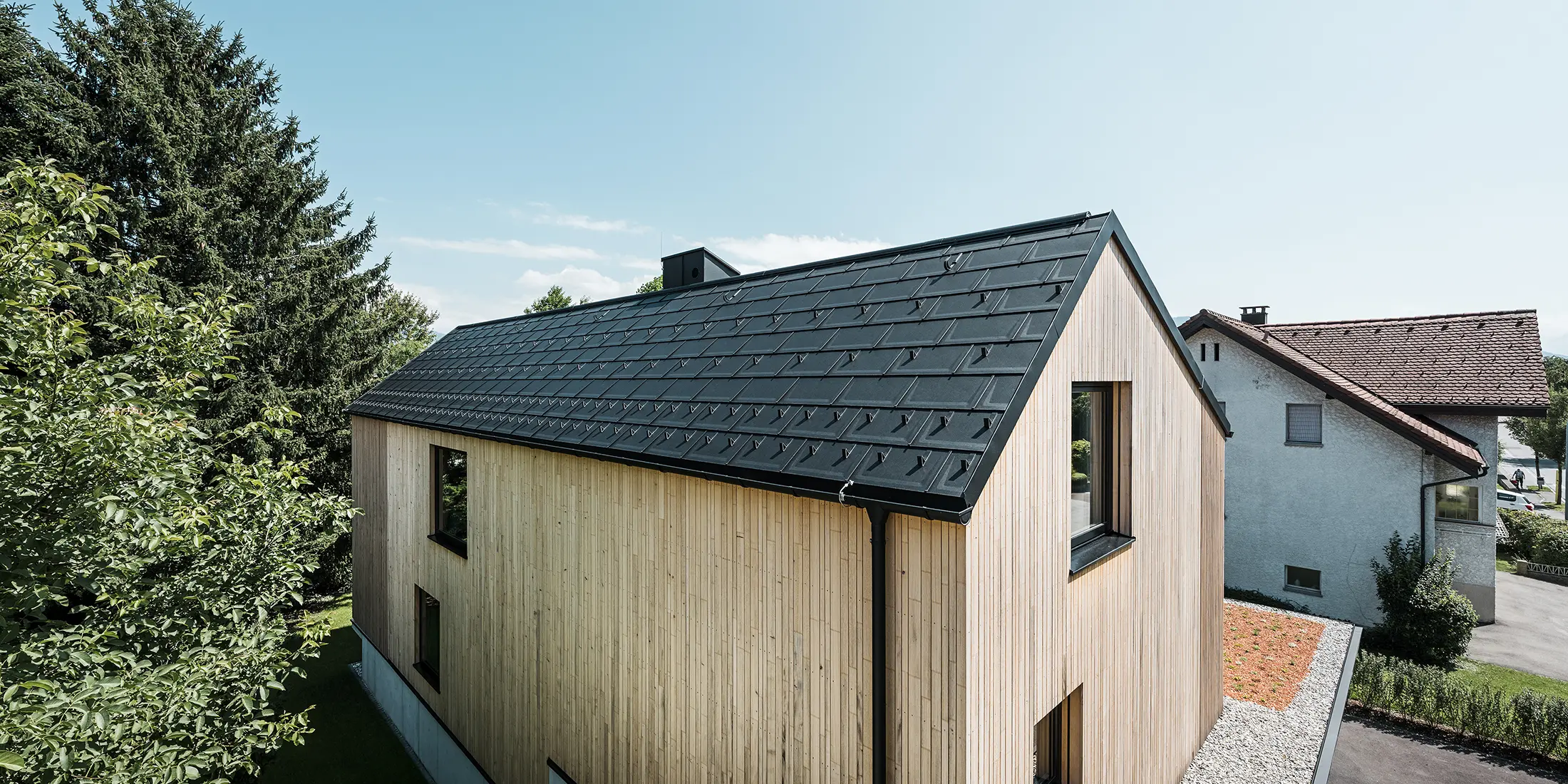 Ein modernes Einfamilienhaus in Lustenau besticht durch die Kombination einer freundlichen Holzfassade mit robusten PREFA Aluminiumprodukten. Das Satteldach wird von der PREFA Dachplatte R.16 mit Schneestoppern sowie der passenden Dachentwässerung in Form von Hängerinne und Ablaufrohr geschmückt. Alle Produkte sind einheitlich im Farbton Schwarz P.10 gehalten und harmonieren perfekt mit der vertikalen Holzfassade.