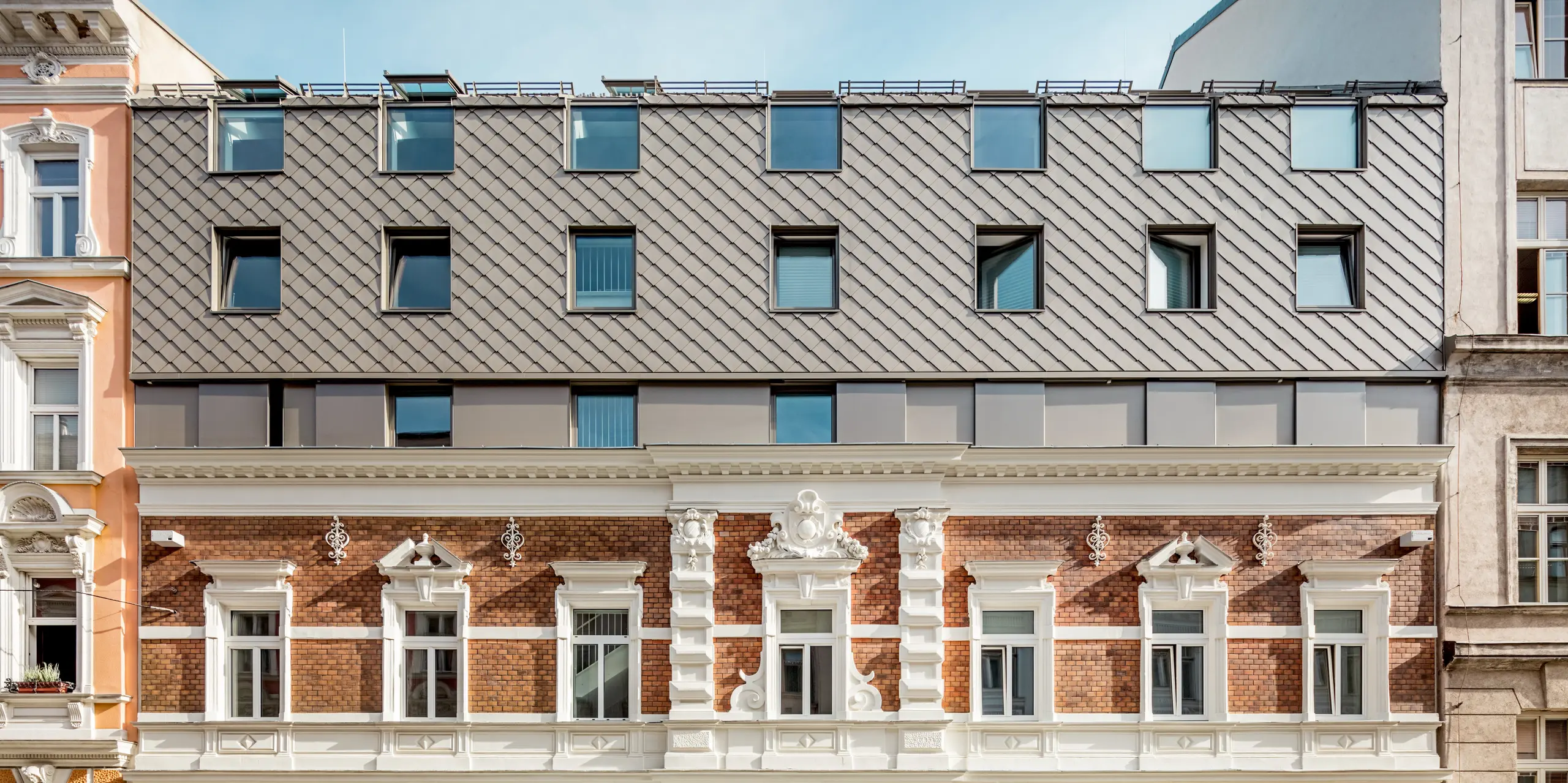 Vue impressionnante de la façade d'un immeuble résidentiel à Vienne, récemment agrandi. L'étage supérieur est revêtu de losanges de façade et de toiture PREFA couleur bronze chaud, qui s'intègre harmonieusement à l'architecture des bâtiments historiques voisins. Les lignes épurées des fenêtres modernes et la structure du bardage, évoquant les tuiles traditionnelles tout en y apportant une touche contemporaine, sont particulièrement remarquables. Cette alliance réussie d'ancien et de moderne confère au bâtiment une identité visuelle unique.
