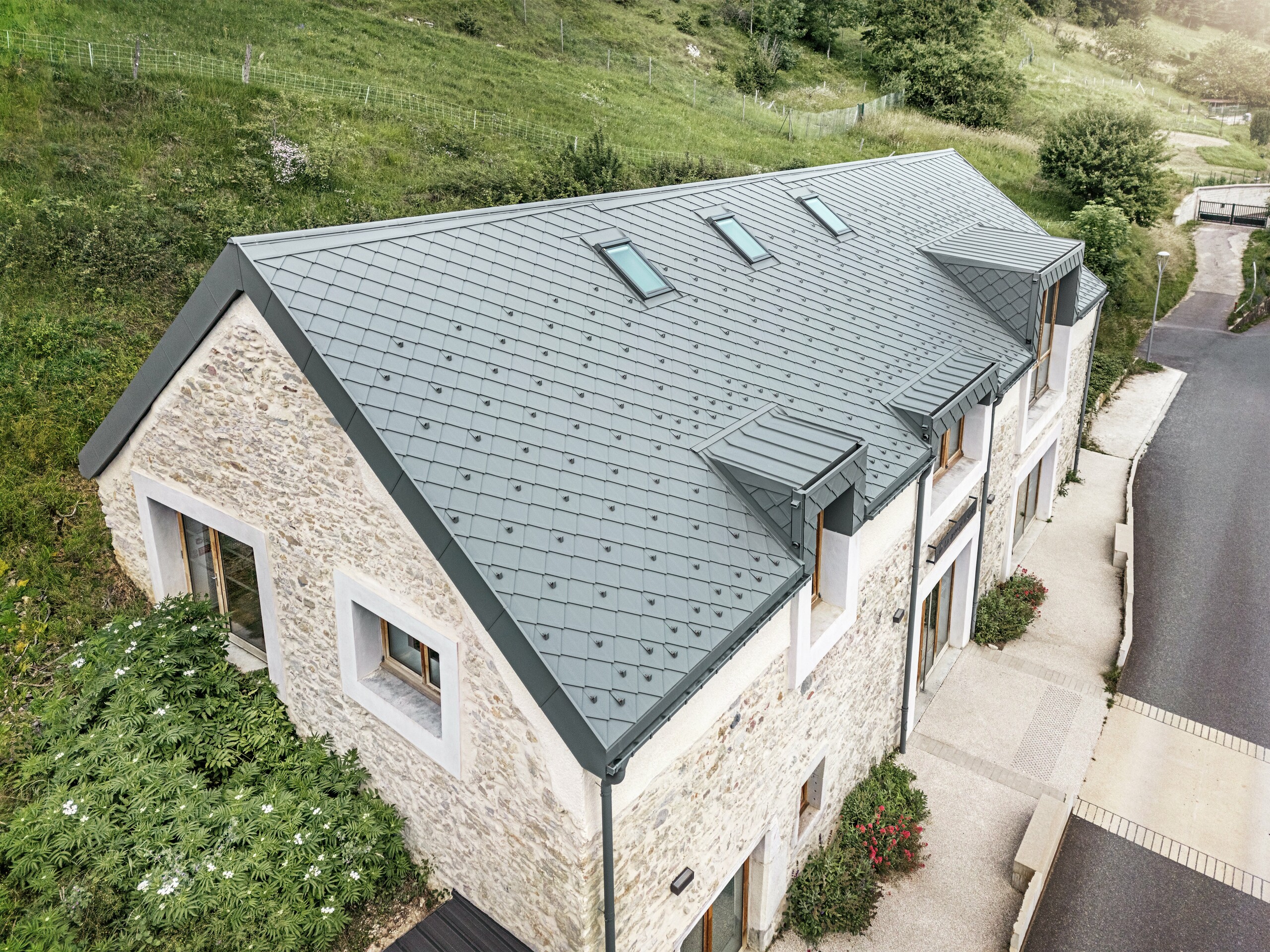 Vue du toit de la grange restaurée "La Grange à Gaby" à Engins, France. Le toit moderne en aluminium PREFA avec des losanges de toiture 29 × 29 s'intègre harmonieusement dans le bâtiment historique en pierre et offre une protection durable contre les éléments.