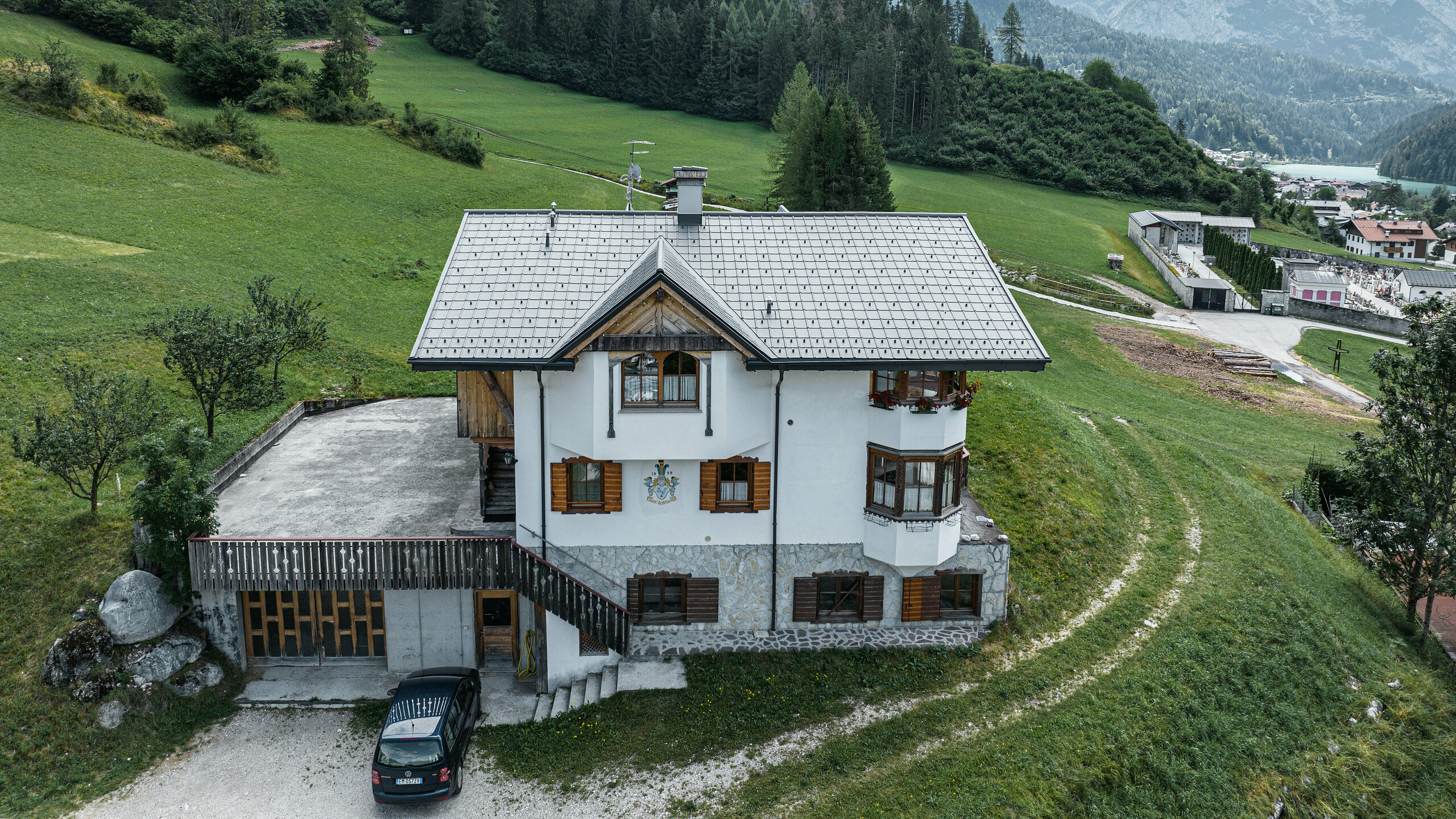 Vue aérienne de l'entrée du refuge de montagne traditionnel d'Auronzo di Cadore après la rénovation du toit avec des tuiles PREFA en aluminium P.10 gris souris. Le bâtiment, avec son charme alpin caractéristique et son toit moderne en tôle, s'intègre harmonieusement dans le paysage verdoyant et le panorama montagneux impressionnant en arrière-plan. Il démontre une combinaison réussie entre fonctionnalité et architecture régionale.