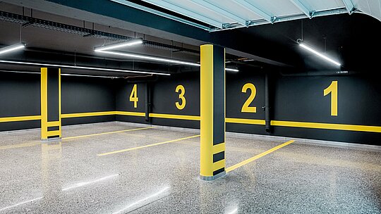 La photographie présente le parking souterrain moderne d'une villa multigénérationnelle à Budapest, conçu dans un style industriel épuré avec des murs sombres et un marquage jaune contrastant. Les places de parking numérotées et les lignes précises confèrent structure et harmonie à l'espace. Le sol poli et brillant reflète la lumière des luminaires LED fins et souligne la qualité des finitions. Le parking s'intègre parfaitement à l'architecture sophistiquée du bâtiment, dont l'extérieur est caractérisé par des produits en aluminium PREFA durables et résistants aux intempéries.