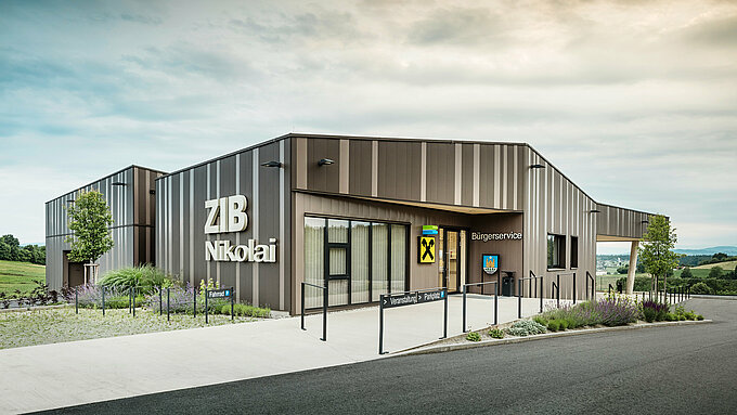 Vue de face du centre événementiel ZIB Nikolai à St. Veit, avec une façade moderne en aluminium à rayures marron-beige-gris et une grande inscription « ZIB Nikolai ». La zone d'entrée présente une signalisation claire pour les événements, le stationnement et les vélos, ainsi qu'un service citoyen intégré et une succursale de la Raiffeisenbank. En arrière-plan, des collines verdoyantes et un vaste paysage s’étendent sous un ciel nuageux.