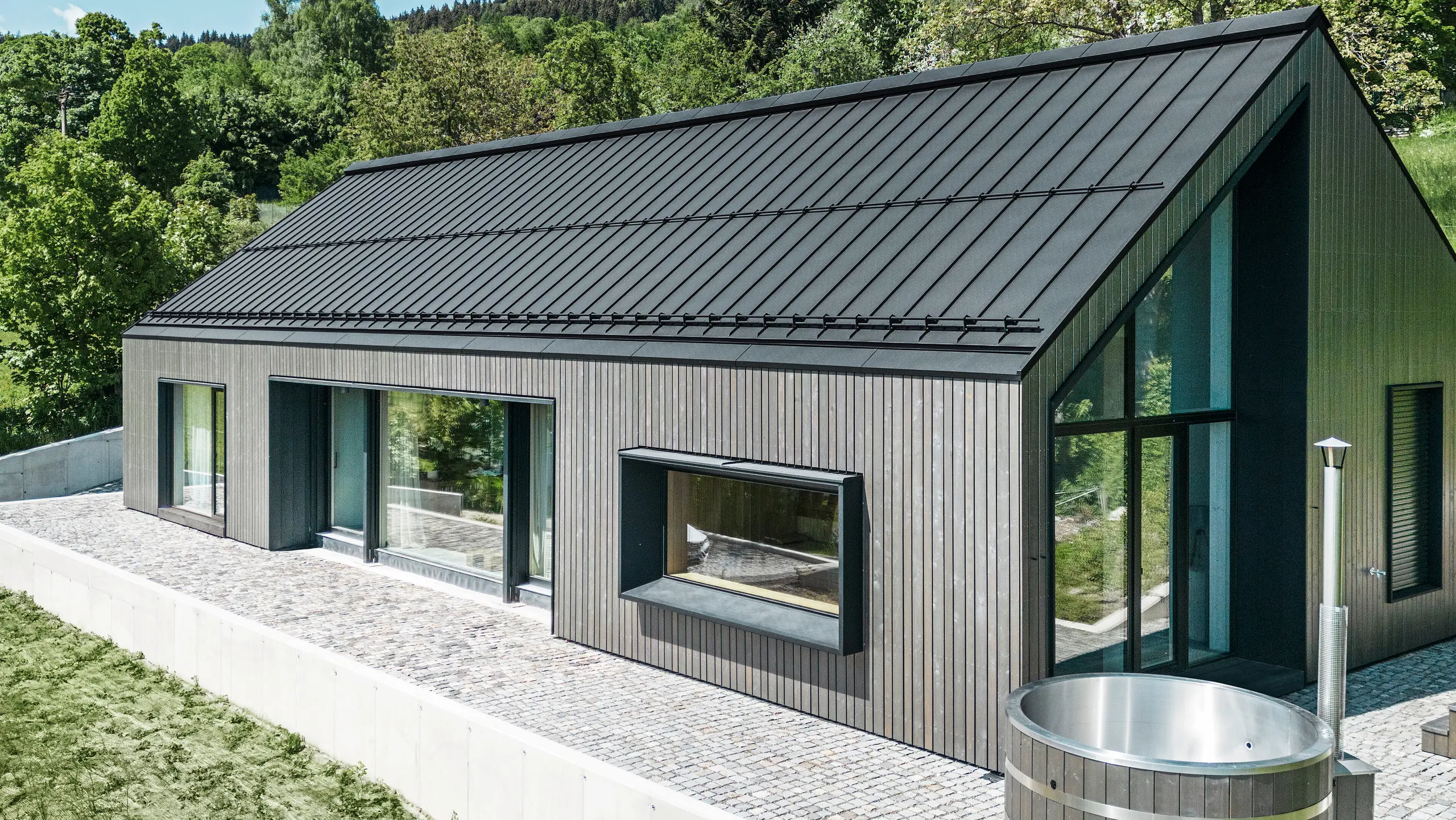 Ein modernes Holzmassivhaus in Kuncice (CZ) wurde mit großen Fensterflächen ausgestattet. Das dunkle Holzmassivhaus verfügt über ein Dachsystem aus PREFALZ in der Farbe P. 10 Schwarz. Im hinteren Außenbereich steht ein Whirlpool mit einer Holzverkleidung.