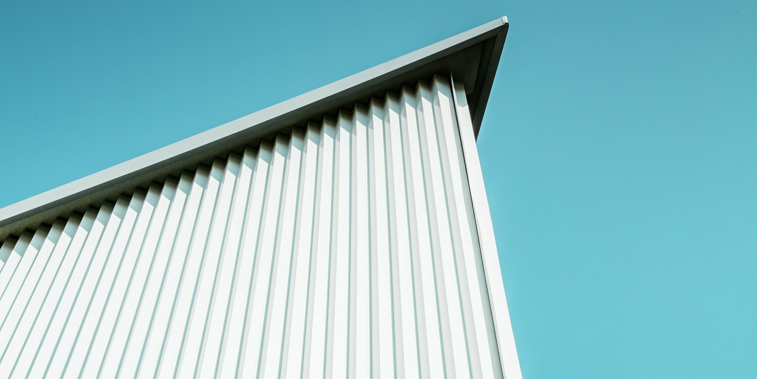 Perspective saisissante de la façade PREFA, avec son profil triangle blanc pur P.10 se détachant sur un ciel dégagé. Les lignes précises du revêtement en aluminium soulignent la netteté architecturale. Résistante aux intempéries et durable, la tôle assure une stabilité maximale et un entretien minimal – idéale pour tous types de bâtiments.