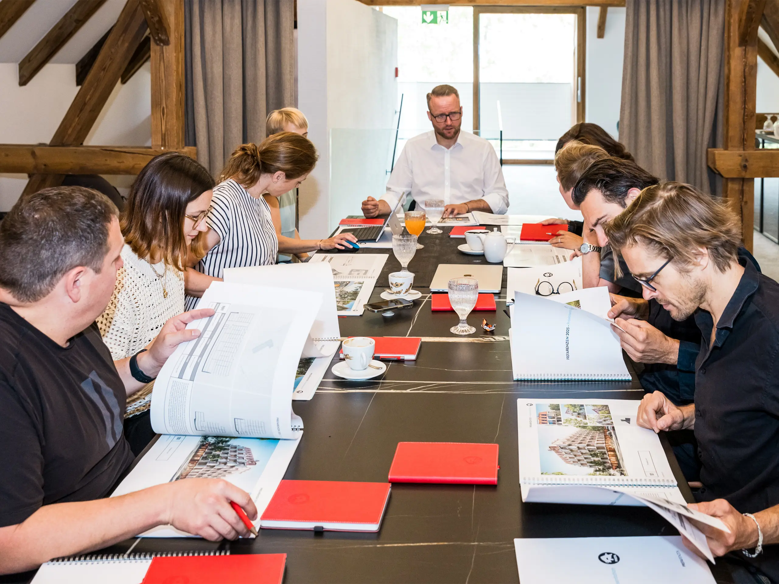 Dialogue d’experts PREFARENZEN 2025 : Dix architectes et experts PREFA sont réunis autour d’une table de conférence dans la résidence d’un vigneron du Seewinkel au Burgenland. Des documents et des plans d’architecture sont étalés sur la table, pendant que les participants débattent avec intensité pour sélectionner les douze projets architecturaux les plus captivants pour la plateforme d’architecture PREFA 2025. La discussion s’est penchée sur les défis écologiques et sociaux, ainsi que sur la fonctionnalité, la simplicité et la valeur ajoutée de l’architecture. Des tasses à café, des verres et des carnets rouges éparpillés sur la table renforcent l’ambiance de travail intense qui y règne.