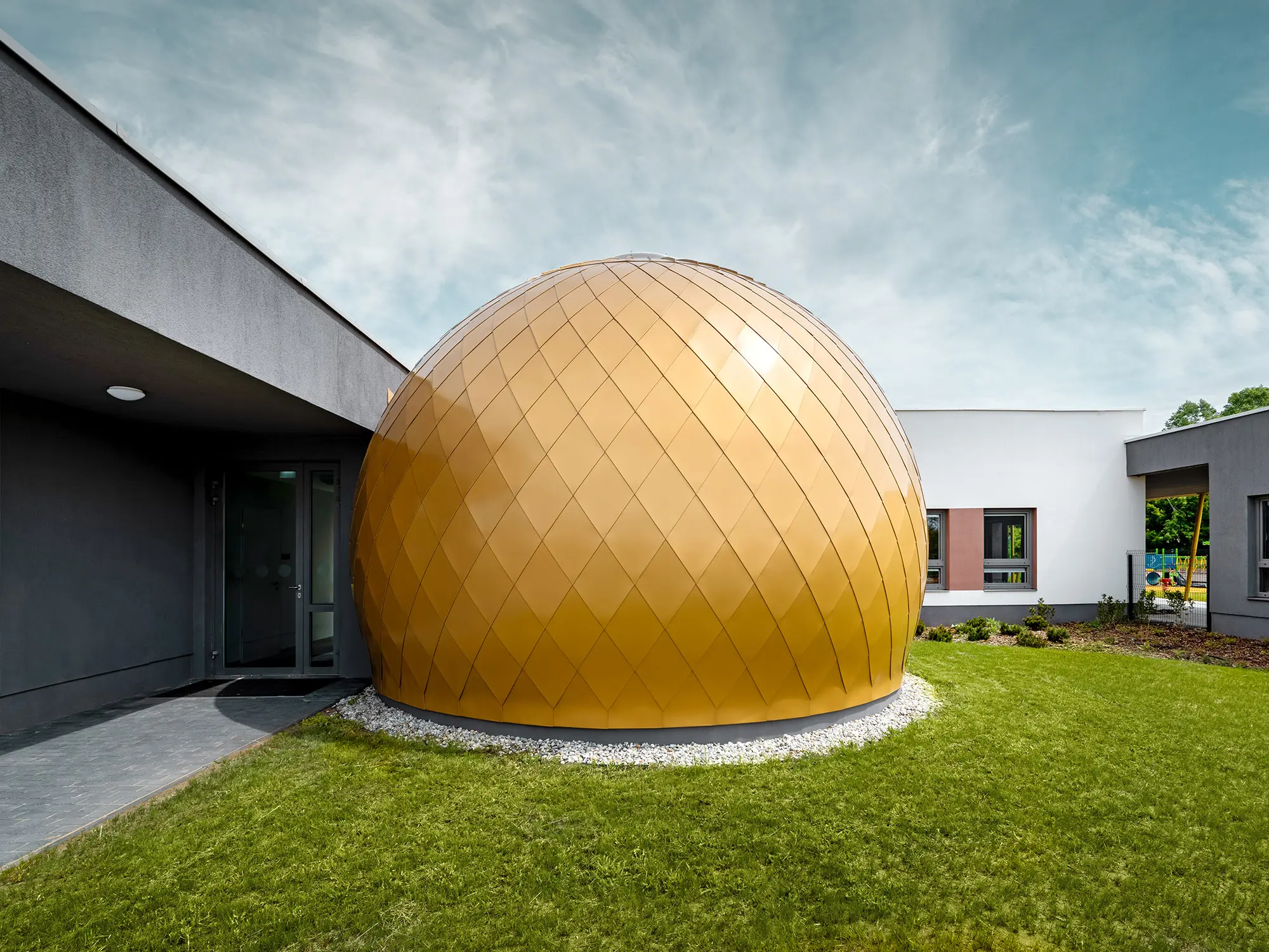 L’une des trois pièces rondes de la crèche conçues par László Erdei vue de côté, derrière et à côté la façade en crépi du bâtiment ainsi que l’aire de jeux sont en partie visibles.