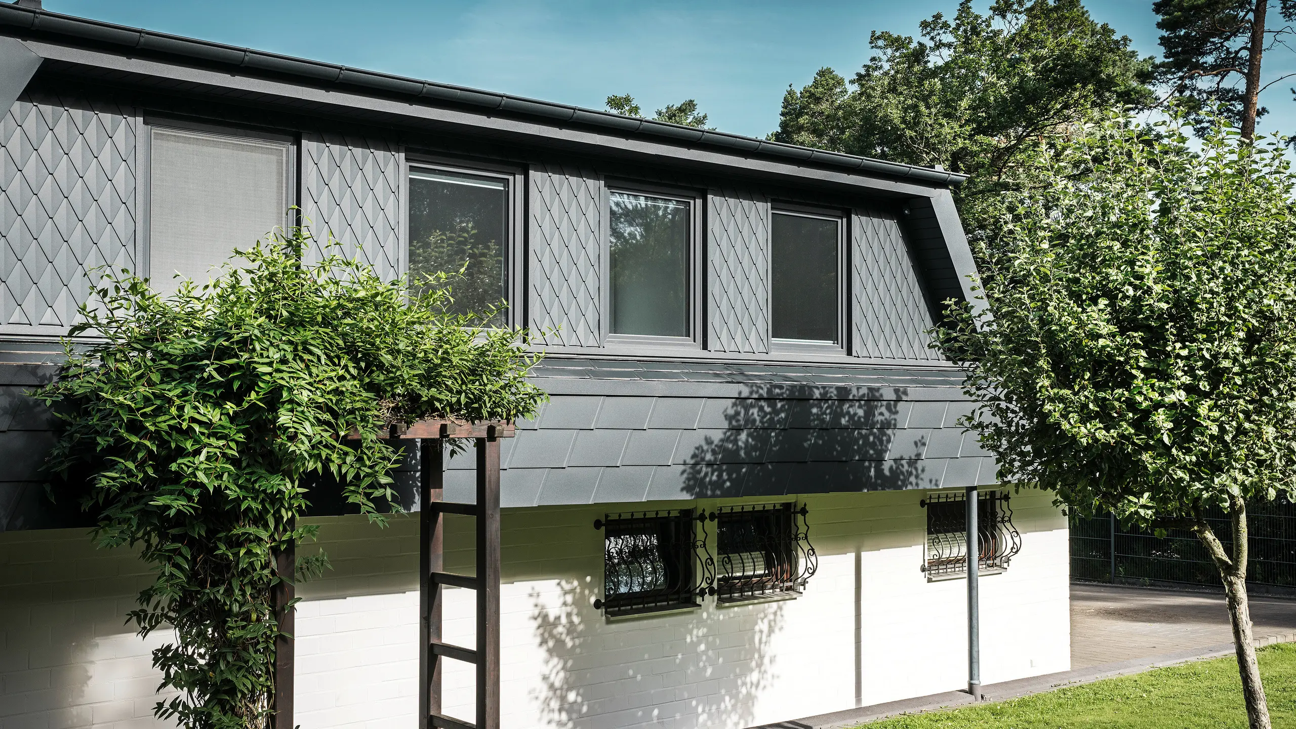 Vue latérale d'une maison individuelle à Mechernich avec de grandes lucarnes trapézoïdales. Les lucarnes sont équipées de petits losanges PREFA en P.10 anthracite, tandis que le toit est recouvert des DS.19 PREFA, également en P.10 anthracite, qui ont fait leurs preuves. Les losanges et bardeaux de toiture en aluminium offrent une solution résistante aux intempéries, esthétique et durable pour la toiture et la façade. La maison unifamiliale traditionnelle est entourée d'un jardin bien entretenu et de nombreux arbres.