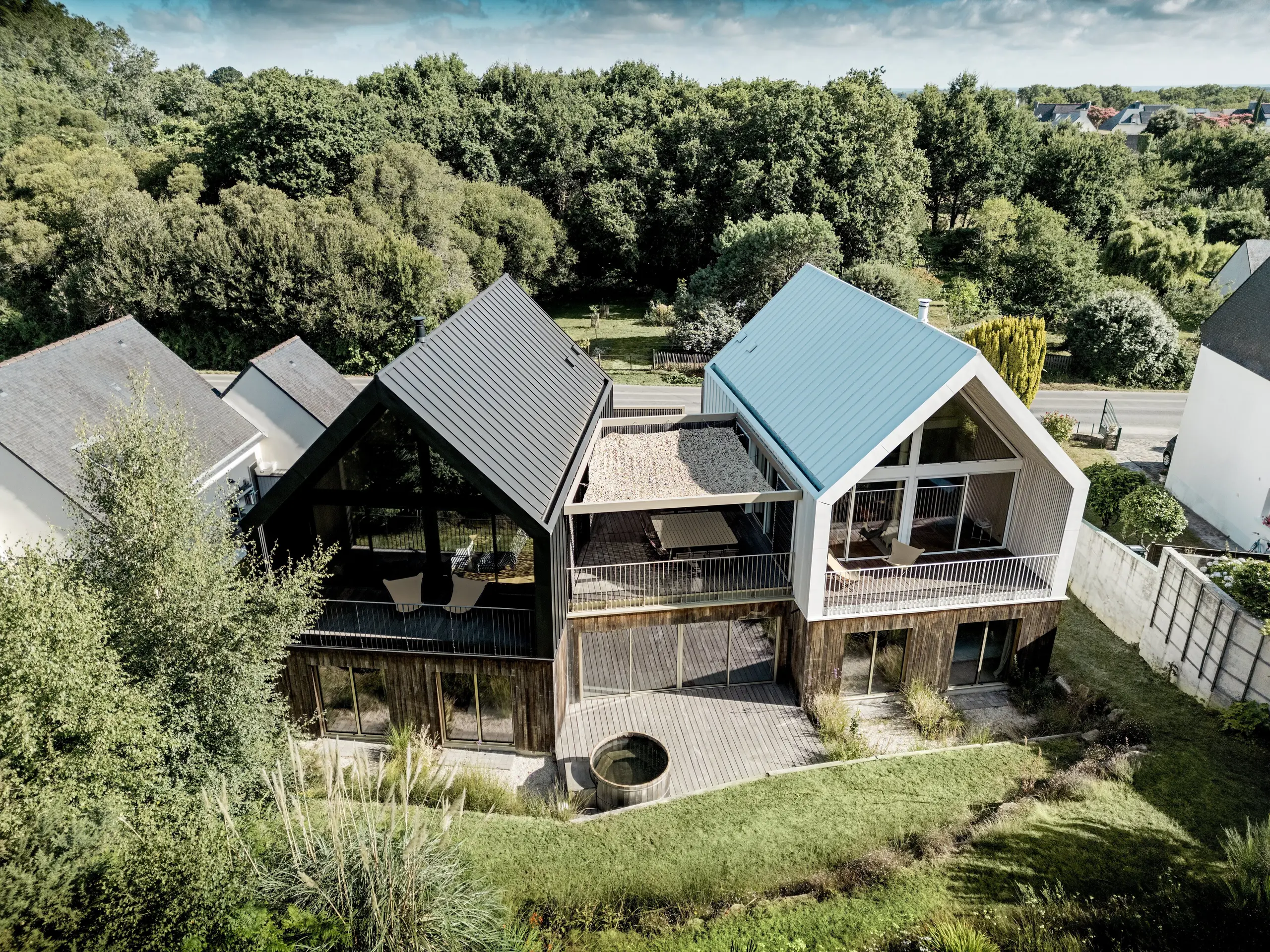 La photographie aérienne du complexe résidentiel Le Rhune révèle deux bâtiments modernes, évoquant les maisons de pêcheurs bretonnes, avec un bardage en aluminium PREFALZ contrasté, en blanc pur P.10 et gris-noir. Les toitures à deux pans remarquables, les larges baies vitrées et la terrasse commune qui les relie forment un point focal architectural fort au cœur d'un paysage verdoyant. Les joints debout continus, assurant une liaison parfaite entre la toiture et la façade, sont particulièrement impressionnants. L'aluminium PREFALZ, durable et résistant aux intempéries, souligne la pureté des lignes, permet une grande précision dans les détails et confère au complexe une allure moderne et haut de gamme.
