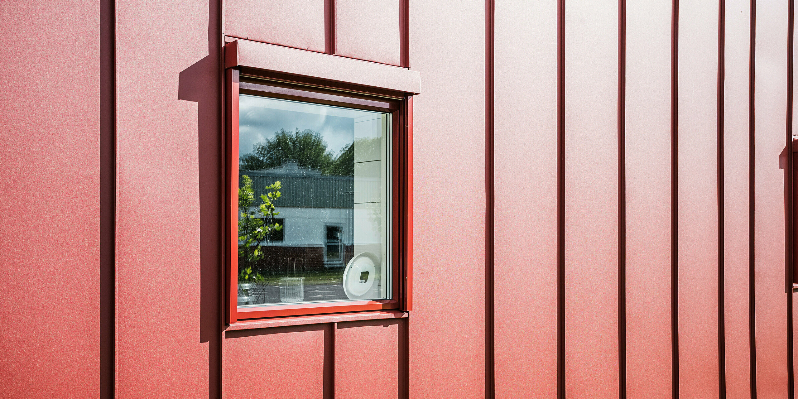 Gros plan d'une fenêtre de la cantine scolaire Gröna Hjärtat à Broby, en Suède, montrant la façade détaillée PREFALZ en rouge oxyde. Le traitement précis de la toiture en aluminium à joints debout souligne la haute qualité et la longévité de l’enveloppe du bâtiment. La fenêtre s'intègre harmonieusement à la façade et reflète l'environnement, ce qui souligne l'architecture moderne et claire du bâtiment. La conception attrayante fait du bâtiment un exemple de construction moderne et durable.