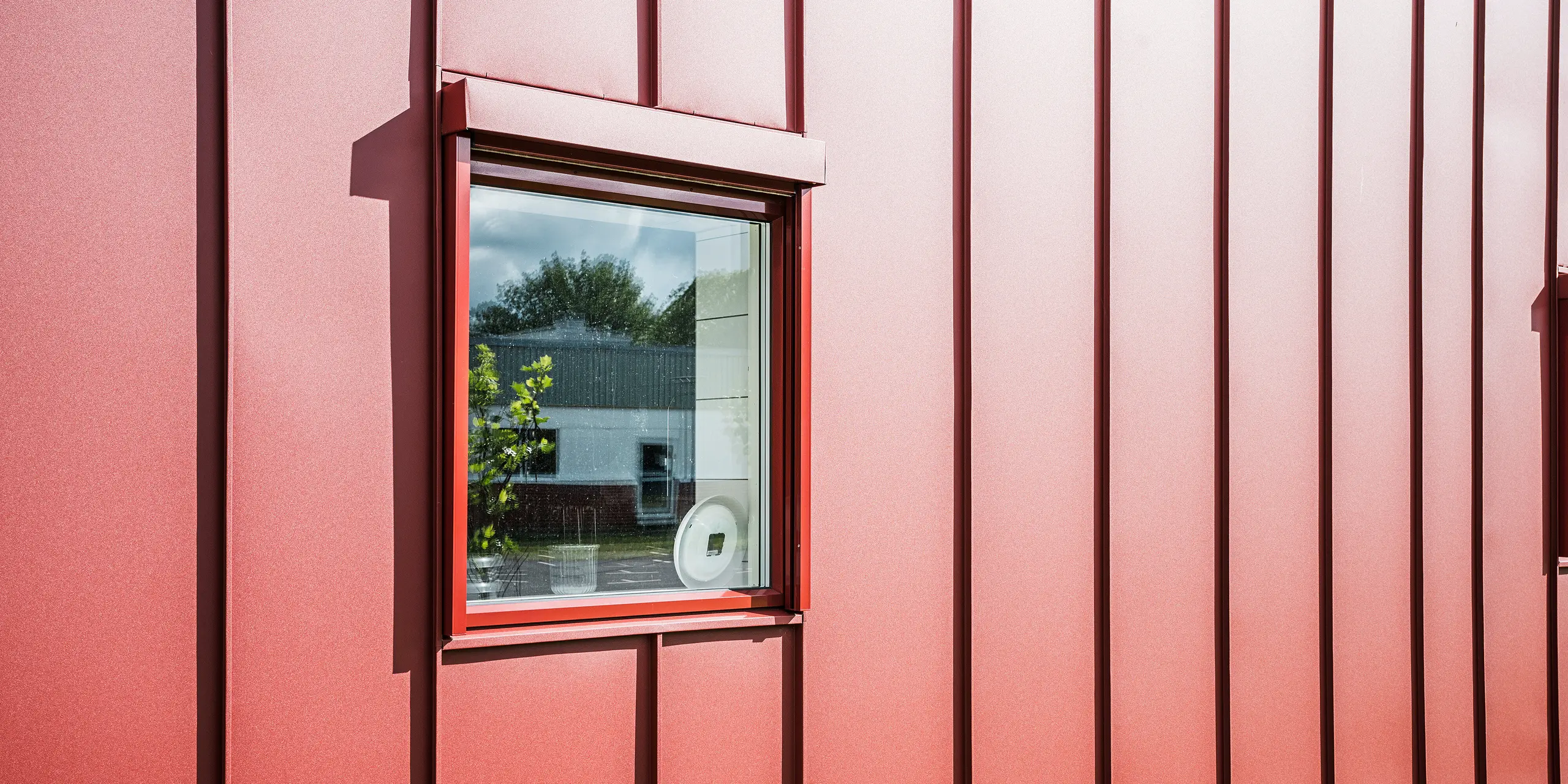 Gros plan d'une fenêtre de la cantine scolaire Gröna Hjärtat à Broby, en Suède, montrant la façade détaillée PREFALZ en rouge oxyde. Le traitement précis de la toiture en aluminium à joints debout souligne la haute qualité et la longévité de l’enveloppe du bâtiment. La fenêtre s'intègre harmonieusement à la façade et reflète l'environnement, ce qui souligne l'architecture moderne et claire du bâtiment. La conception attrayante fait du bâtiment un exemple de construction moderne et durable.