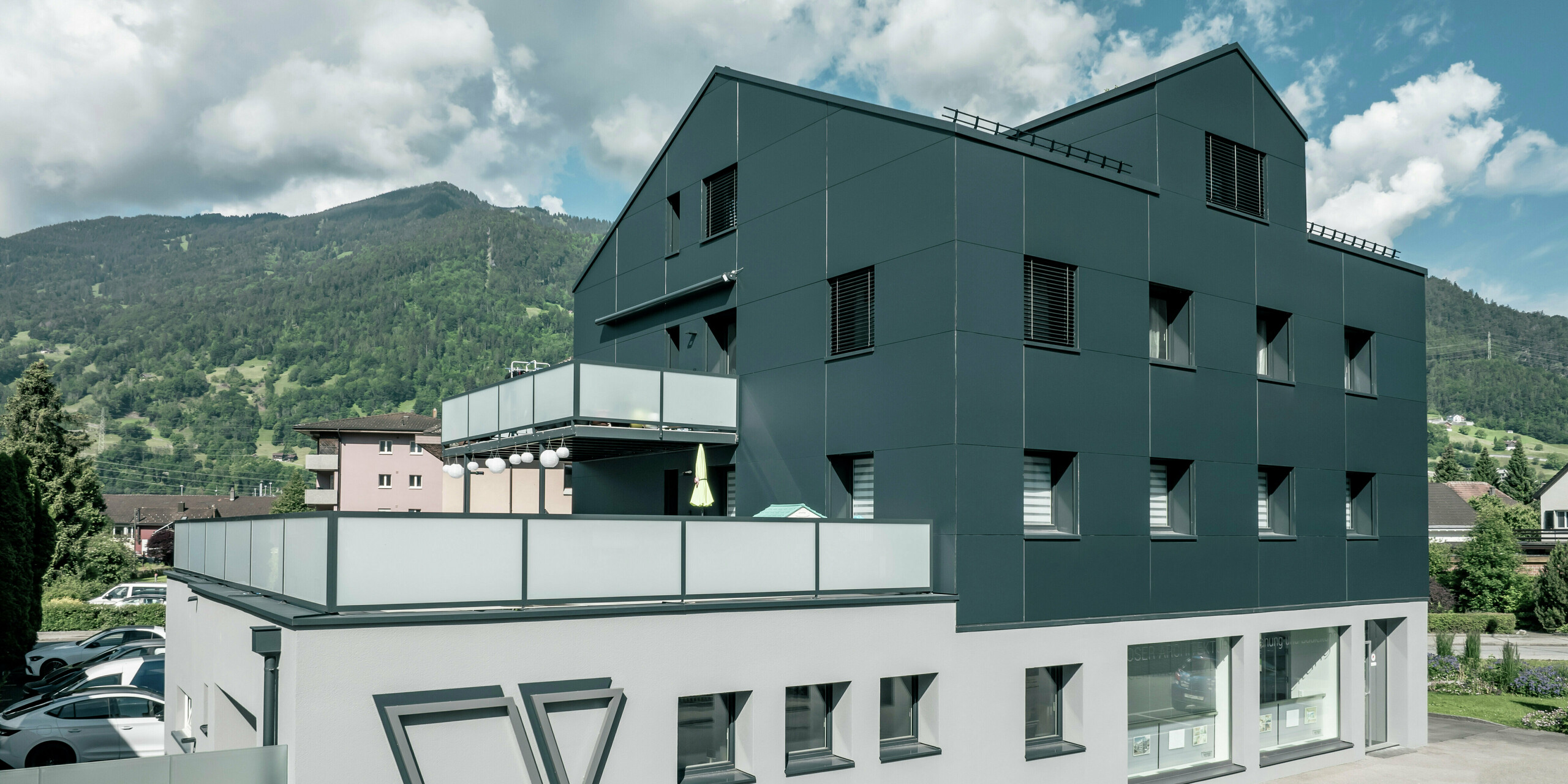 Le bureau d'architecture Voser à Landquart, dans le canton suisse des Grisons, avec une façade en aluminium PREFA en anthracite. Le bâtiment a été rénové et équipé de panneaux composites en aluminium PREFABOND. Le design allie des lignes claires et des matériaux de haute qualité. Les grands balcons avec parapets en verre et le logo frappant du cabinet d'architectes Voser sur le côté du bâtiment sont frappants. Un paysage de montagne verdoyant peut être vu en arrière-plan.