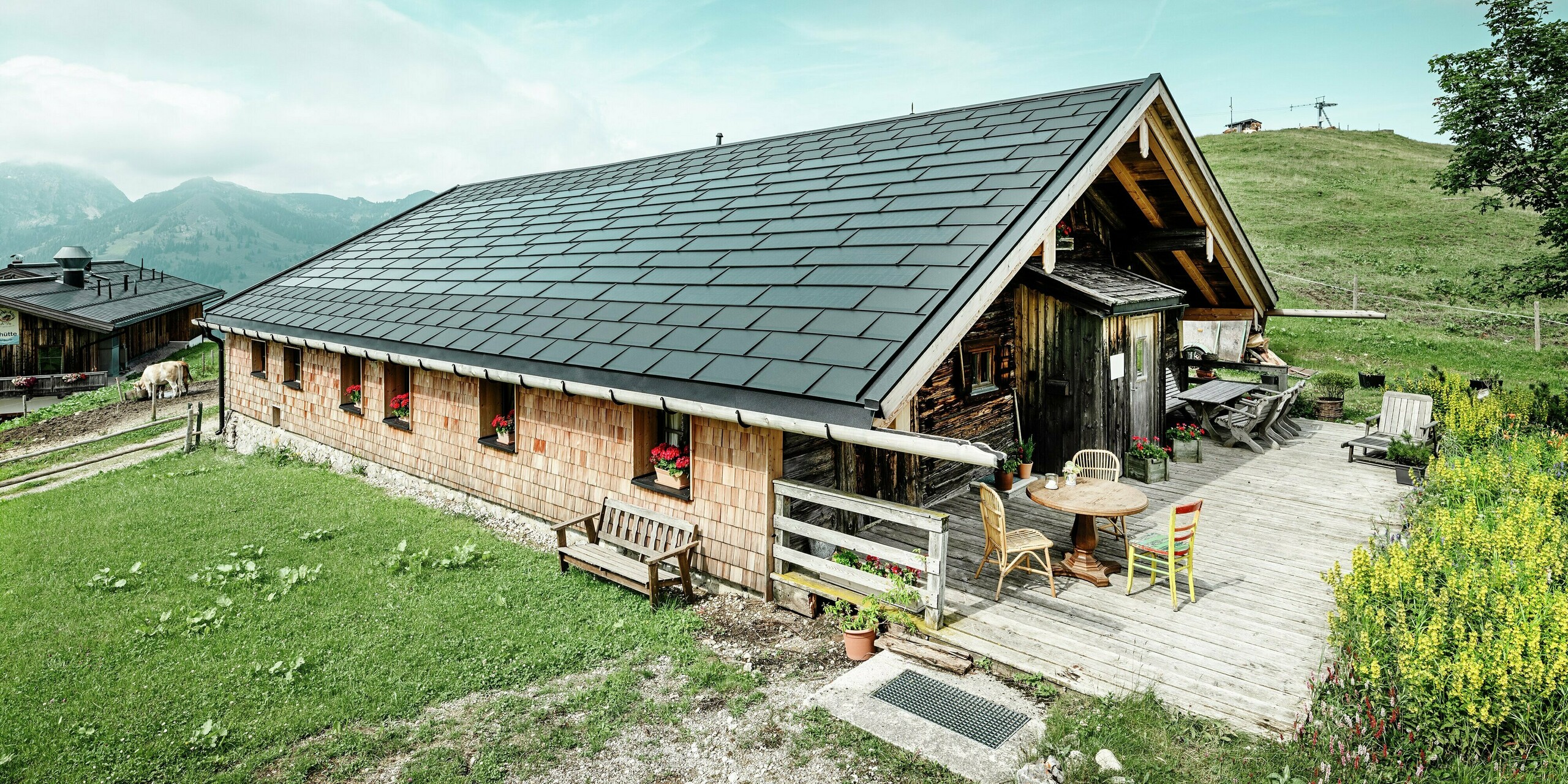 Vue de face de la Birkehütte sur le Walleralm à Bayrischzell, nichée dans le monde pittoresque des montagnes. Lors de la rénovation, le toit a été équipé de tuiles solaires innovantes PREFA. Le système de toiture moderne combine un toit en aluminium durable avec un système photovoltaïque intégré. La surface du toit est complétée par des panneaux de toit PREFA robustes R.16 de couleur P.10 noir. La combinaison de la technologie photovoltaïque moderne et de la construction traditionnelle en bois assure une protection durable contre les intempéries et s'intègre harmonieusement dans l'environnement alpin. Au premier plan de la photo, vous pouvez voir la terrasse accueillante avec des meubles en bois rustique.