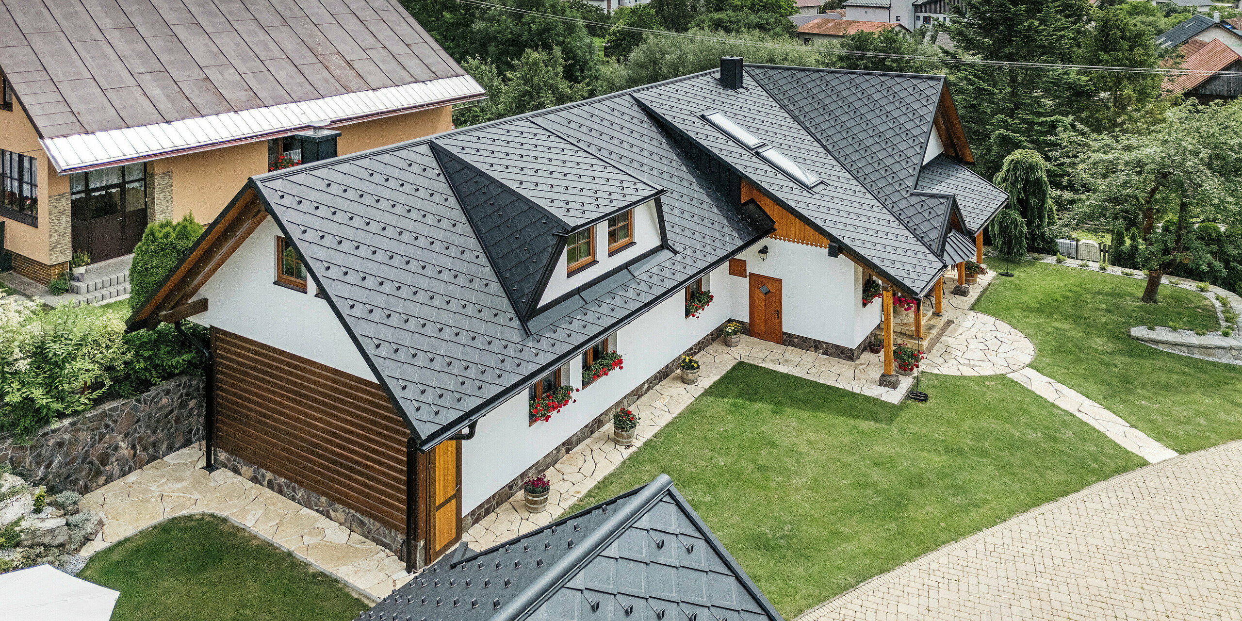 Vue aérienne d'une maison familiale traditionnelle à Stara Bystrica, en Slovaquie, recouverte d'une toiture en losanges PREFA 29 × 29 de couleur P.10 noir. Le toit impressionne par son élégant aspect cabanon et l'intégration harmonieuse des lucarnes. Le paysage verdoyant environnant, le jardin bien entretenu et l'allée pavée soulignent l'aspect général attrayant du bâtiment.