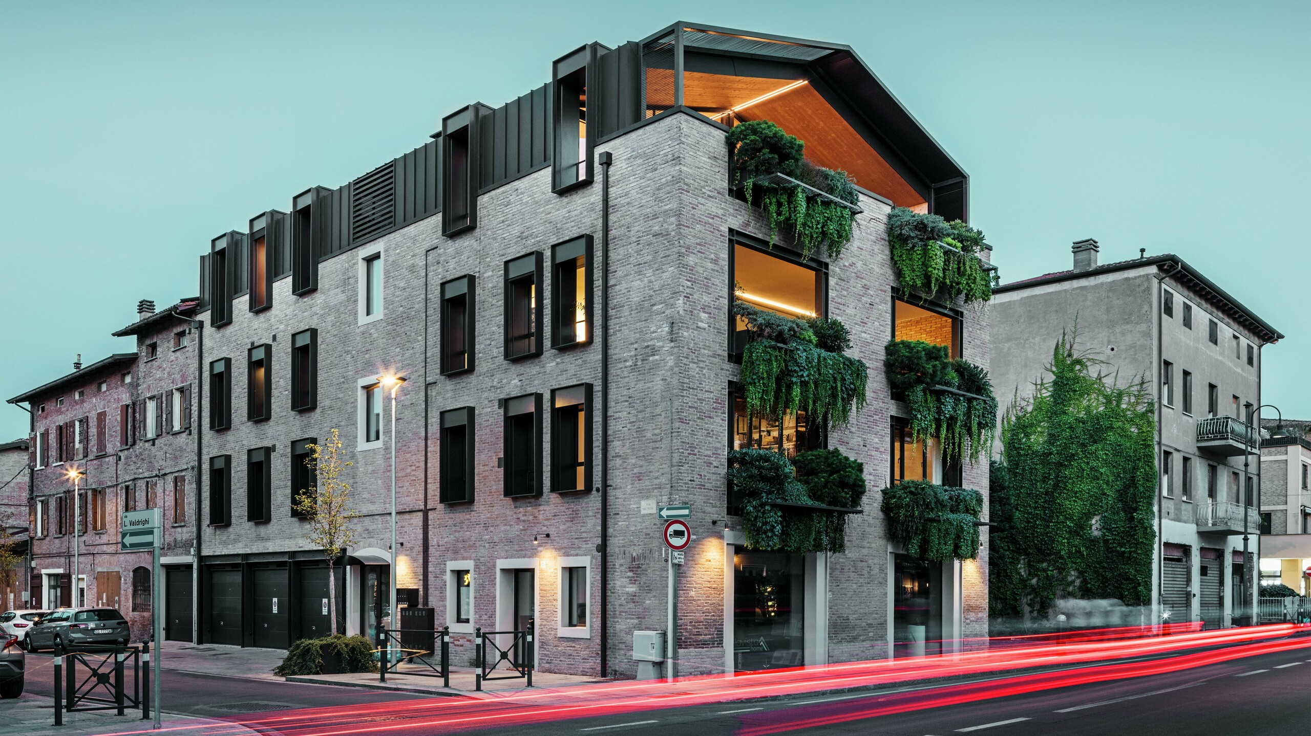 Immeuble résidentiel et commercial durable en Italie avec toiture en aluminium PREFALZ en P.10 brun noisette, façade en briques végétalisée et architecture moderne dans l'ambiance du soir avec des lampadaires lumineux.